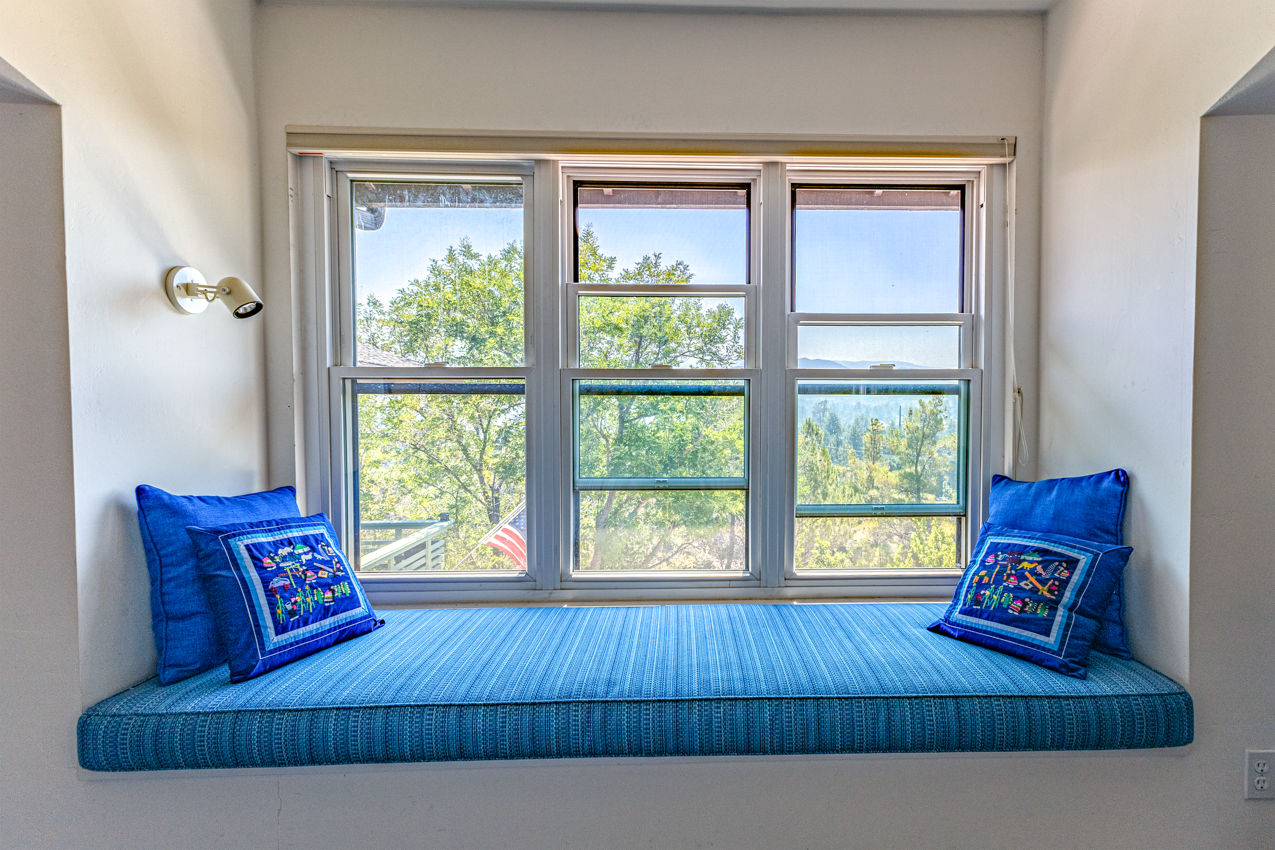 A cozy window seat with blue cushions and embroidered pillows, overlooking a lush green landscape with trees and mountains in the background.