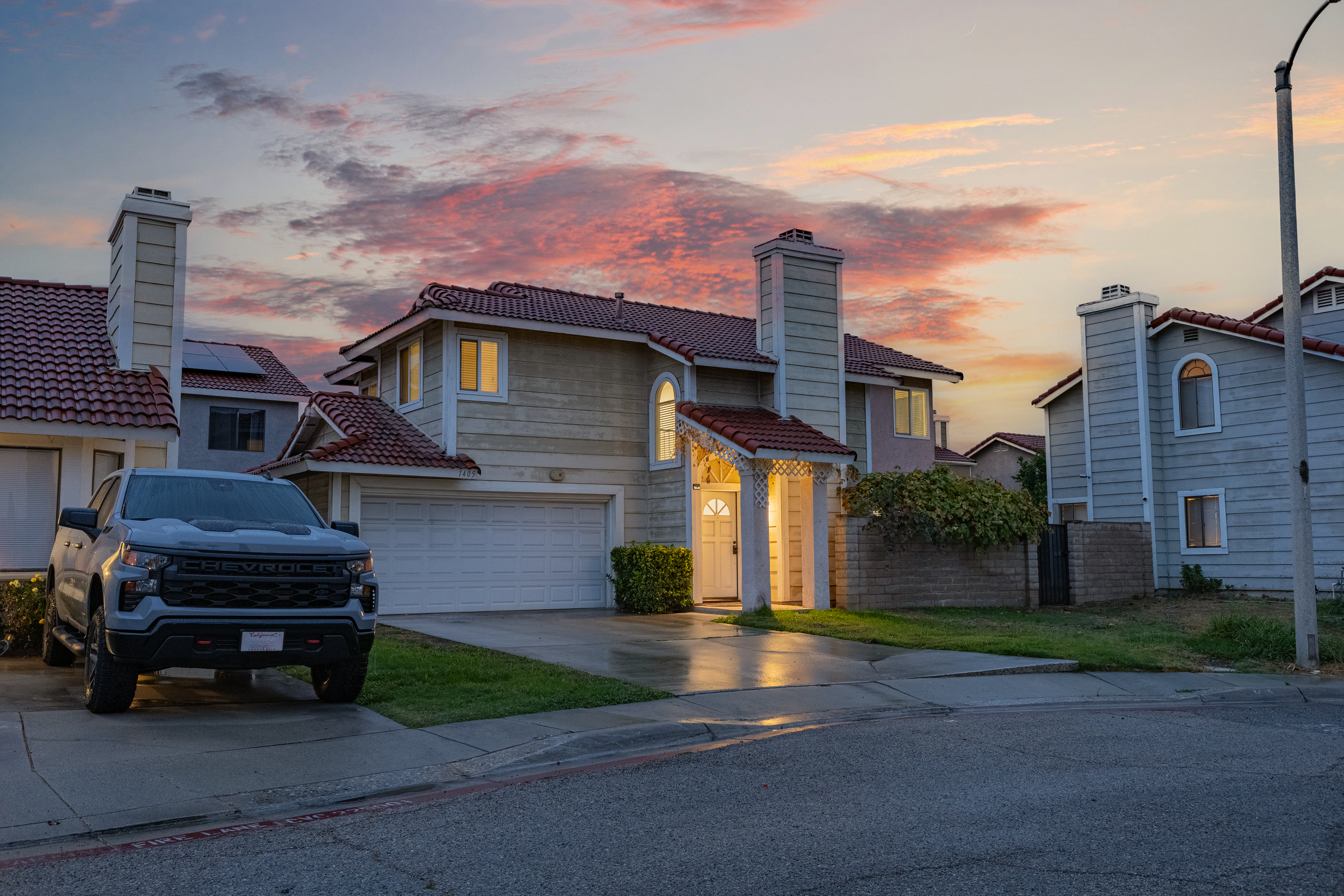A residential street at sunset with a house featuring a white garage door, lit windows, and a front porch. A gray Chevrolet pickup truck is parked in the driveway on the left.
