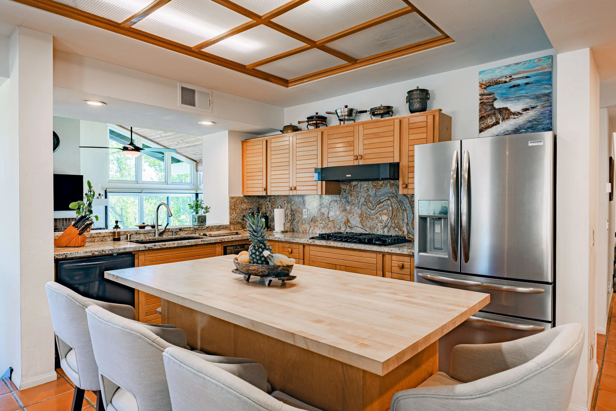 A kitchen with wooden cabinets, granite countertops and backsplash, stainless steel refrigerator, and a wooden kitchen island with four beige chairs. A pineapple and other fruits are on the island. There is a window above the sink, and a painting on