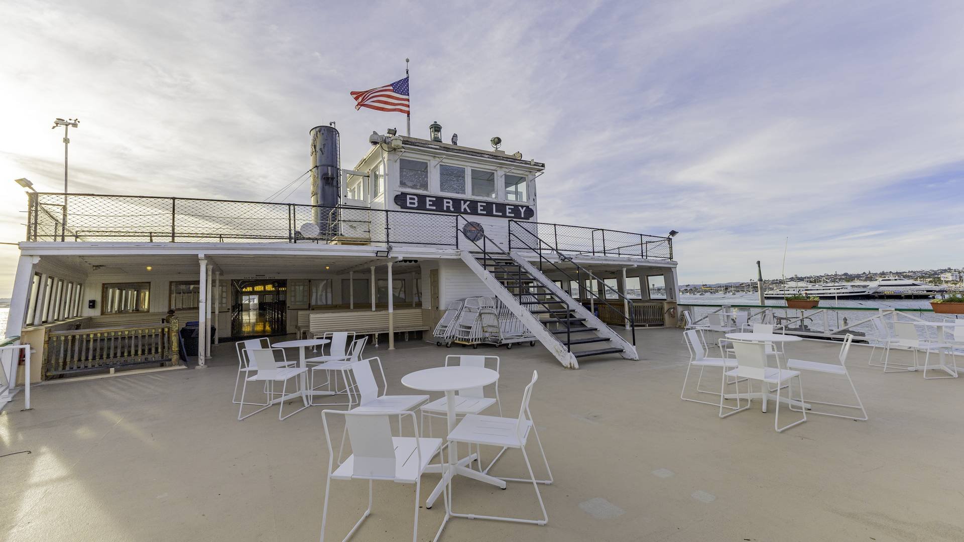 Empty outdoor patio on a boat with white tables and chairs, and a railing overlooking the water, with a Berkeley sign on a boat structure, American flag, and harbor in the background.