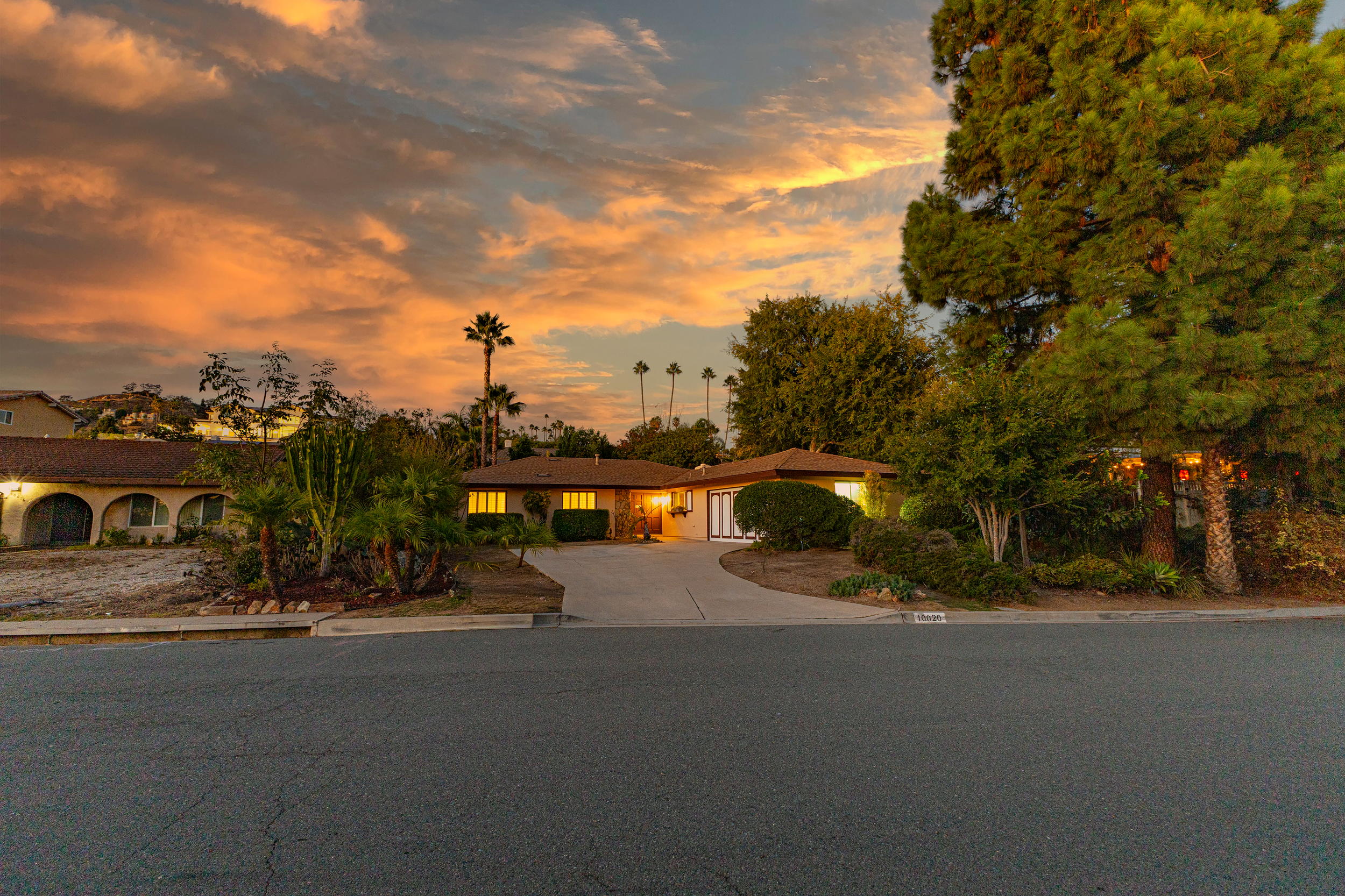 A suburban house is captured at sunset with an illuminated entryway, surrounded by various trees and plants, including palm trees and a large pine tree, with a curved driveway leading to the garage.