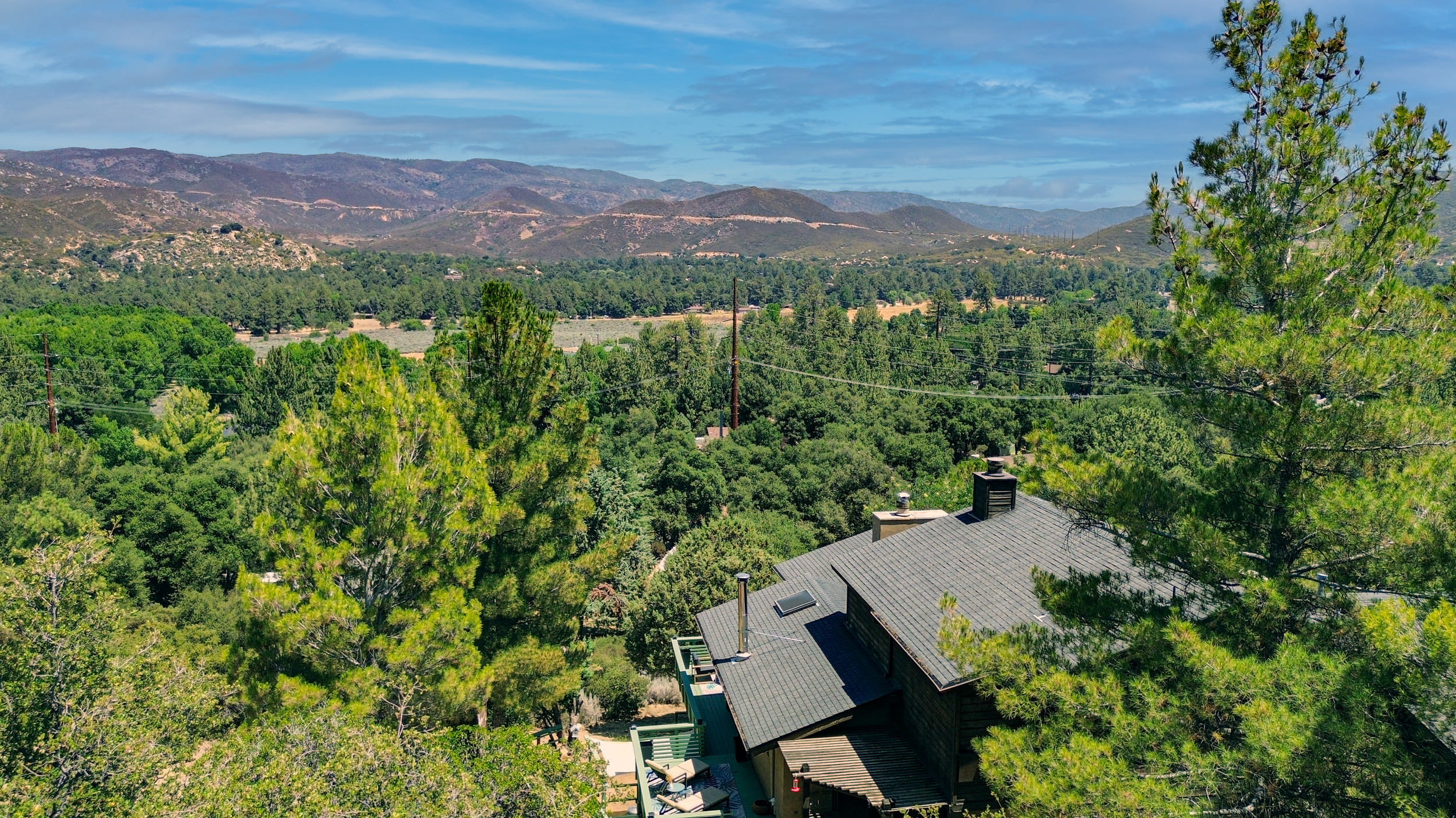 View of a house with a gray roof surrounded by green trees, overlooking a lush valley with hills and mountains in the background under a partly cloudy sky.
