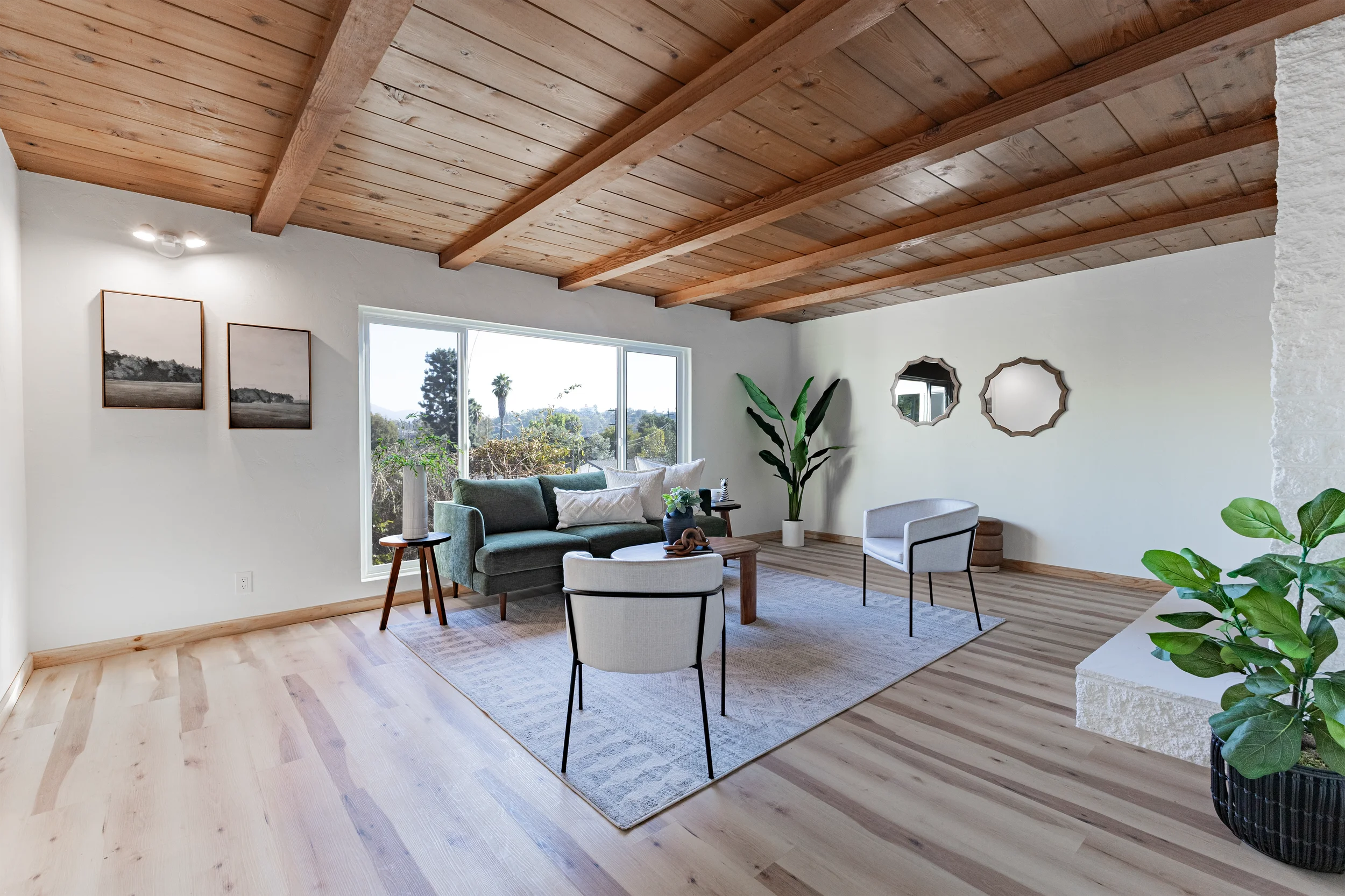 Bright living room with wooden ceiling beams, large window with outdoor trees, green sofa, two white armchairs, potted plants, wall mirrors, and framed artwork.