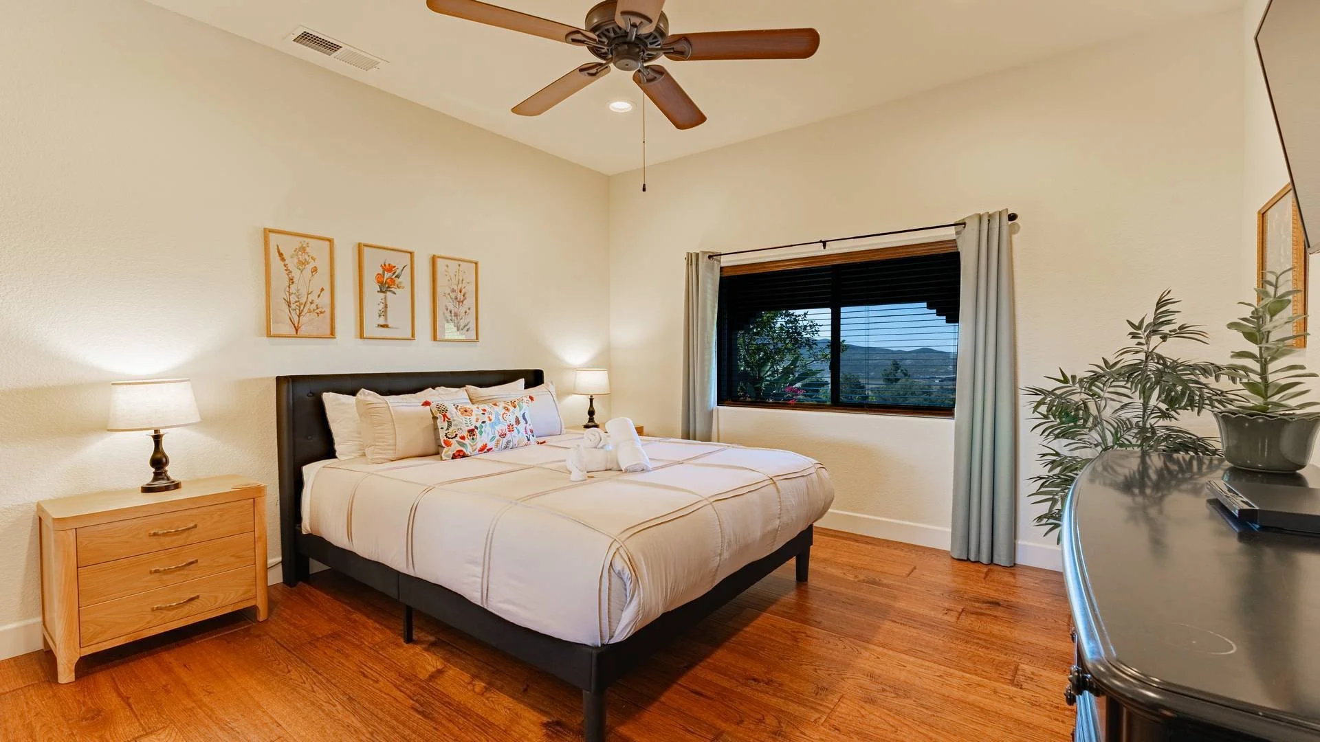 A bedroom with a large bed with white bedding, a black headboard, and colorful pillows. There are two nightstands with lamps, a window with curtains, and framed floral artwork on the wall. A ceiling fan is above, and there are houseplants on a dresse