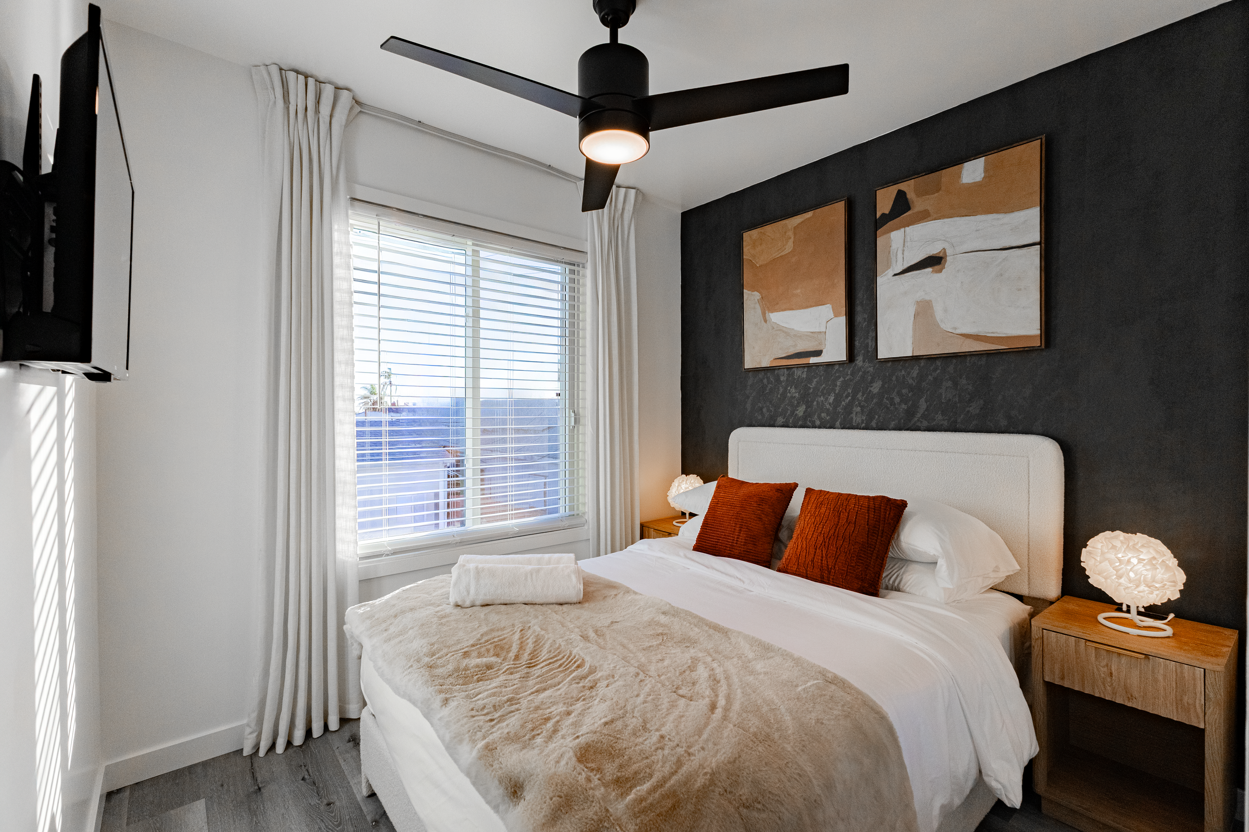 Bright bedroom with a large window, white curtains, black ceiling fan, dark accent wall with two paintings, white bed with rust-colored pillows, beige blanket, fluffy white lamp on wooden nightstand.