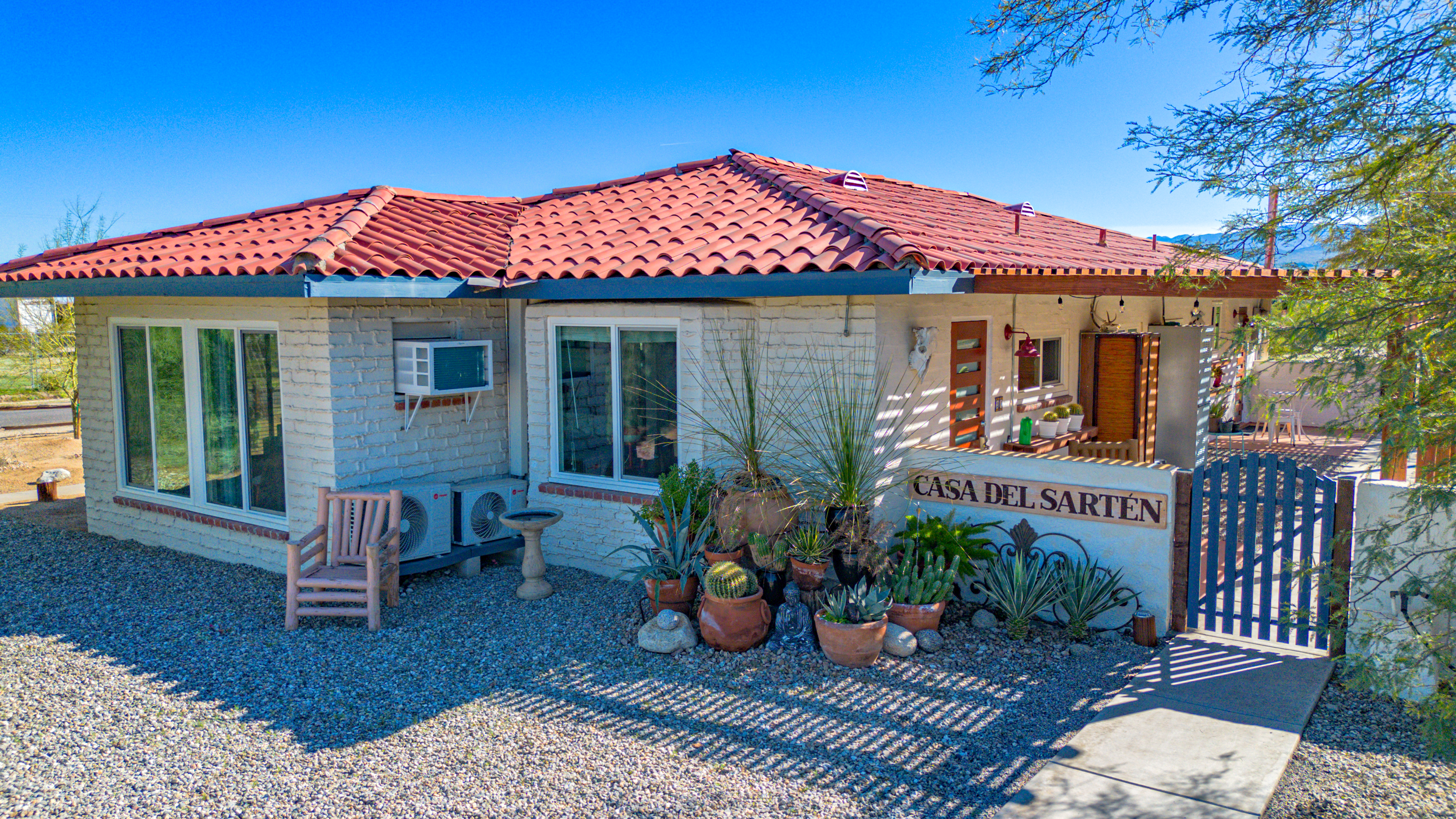 A house with a red tiled roof, white brick exterior, and potted plants outside. There are windows, an air conditioning unit, and a sign that reads 'Casa del Sartén'.