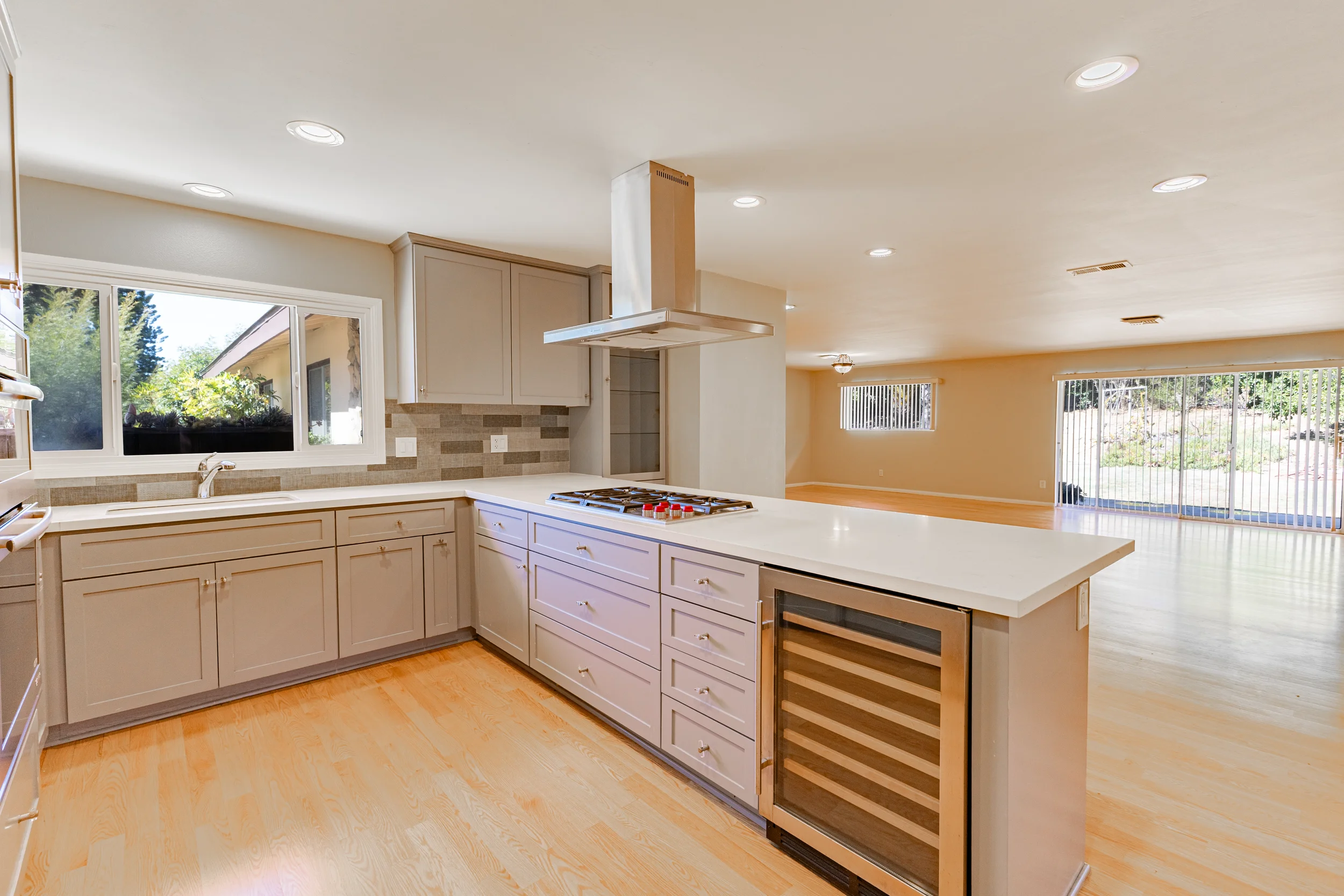 Open-concept kitchen with beige cabinets, a large kitchen island, a stove with a range hood, and a built-in wine cooler. Large windows and sliding glass doors provide natural light and a view of the backyard.