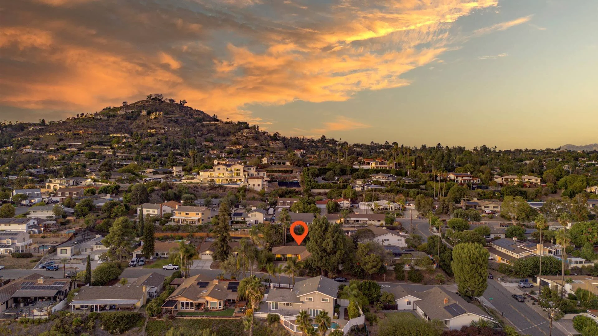 A suburban neighborhood with houses, trees, and a hillside under a partly cloudy sky is shown at sunset.