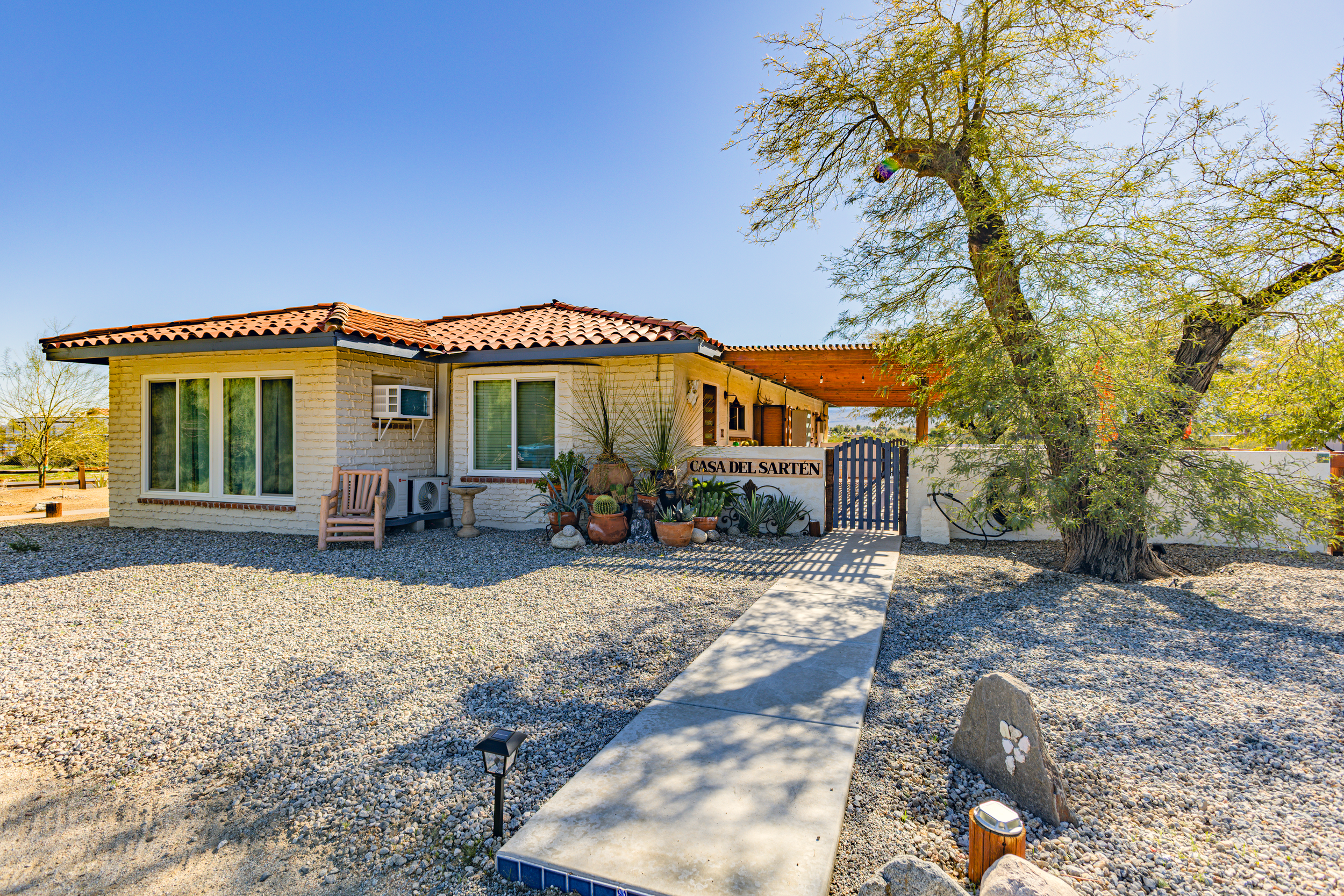 Single-story house with a red tile roof, white brick walls, and large windows, surrounded by desert landscape with gravel ground, a large tree, assorted potted plants, a pathway leading to the entrance, and a blue sky in the background.