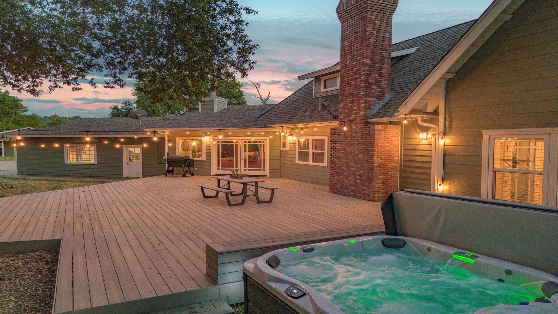 A backyard deck area at sunset with string lights, a hot tub, a picnic table, a grill, and the back of a house with multiple windows and a brick chimney.