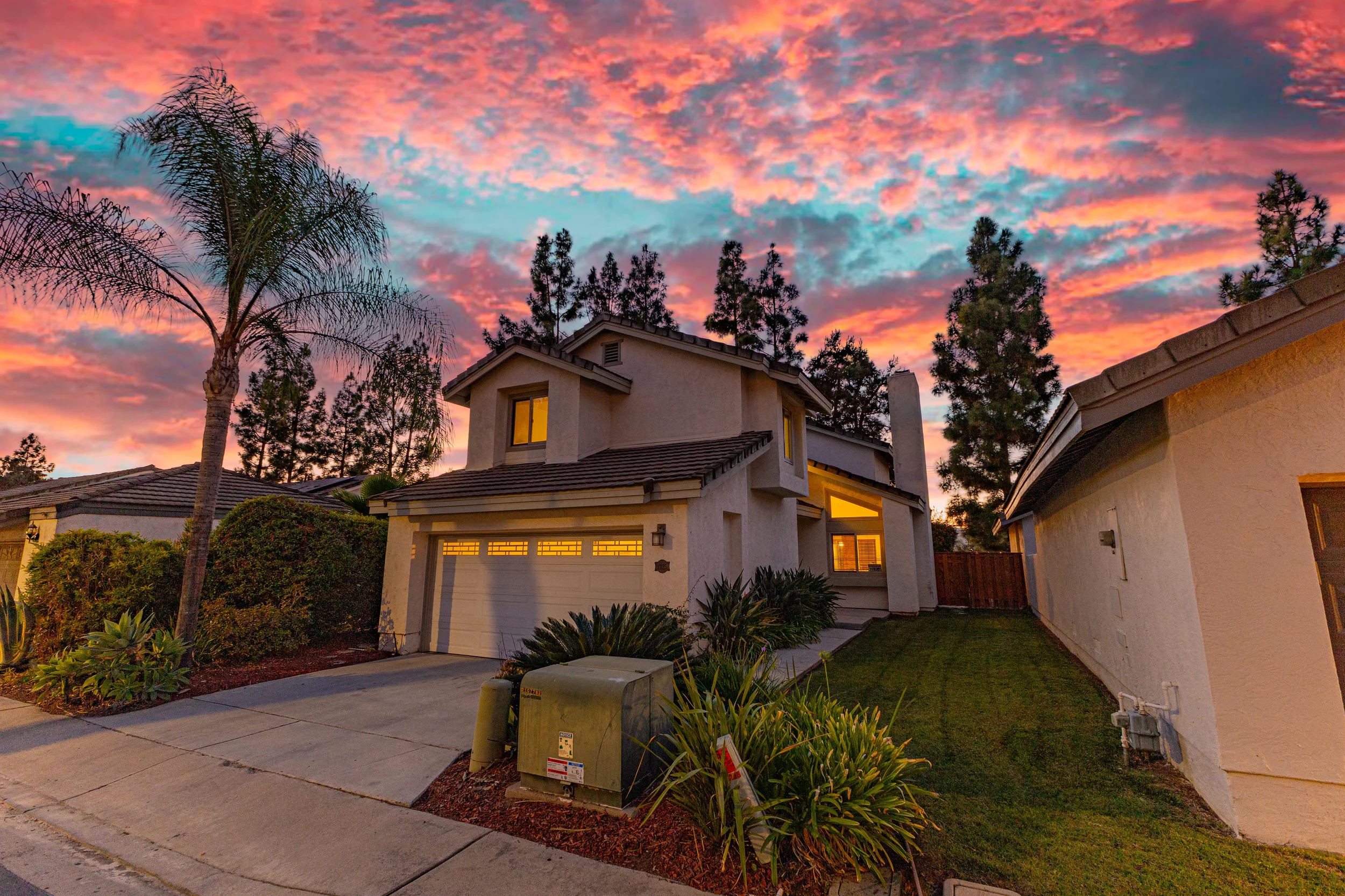 A two-story house with a closed garage door, surrounded by a green lawn, bushes, and tall trees, with a colorful sunset sky in the background.