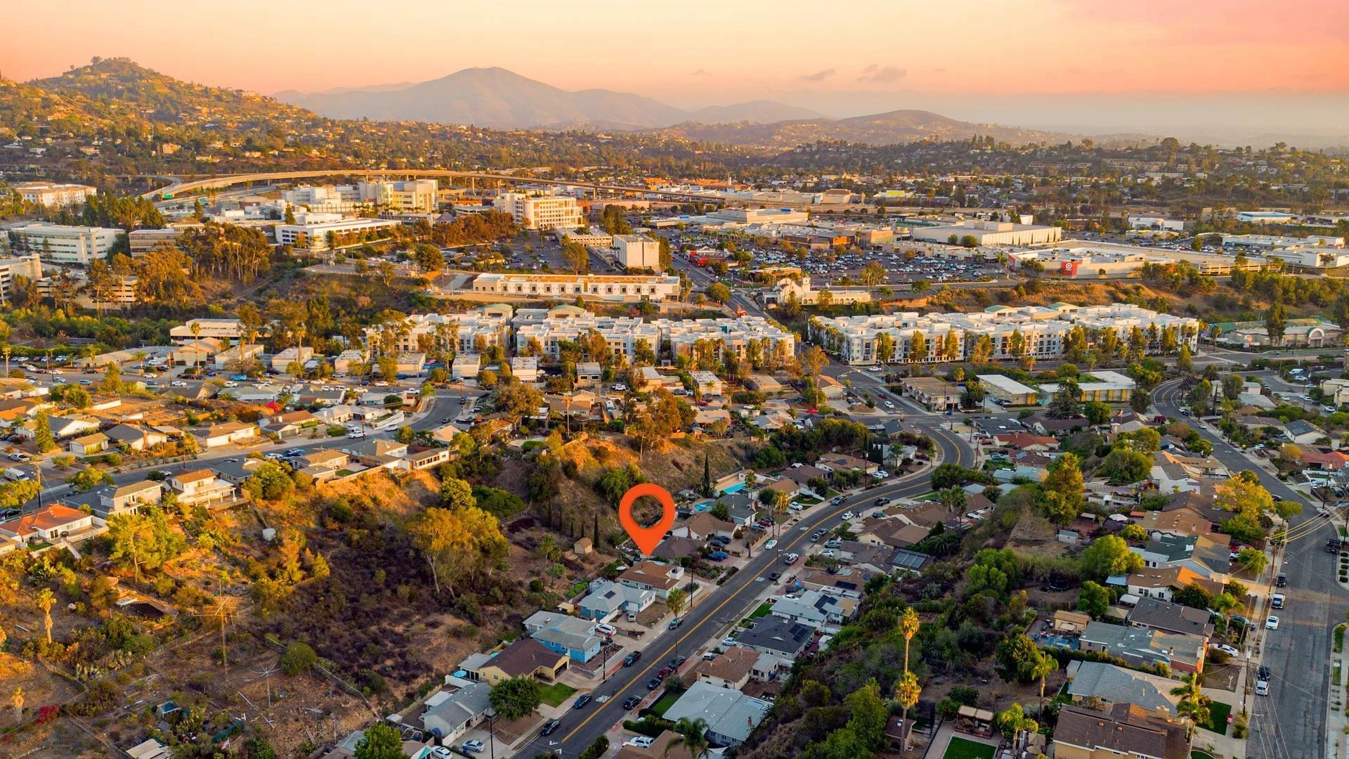 Aerial view of a cityscape at sunset showing residential neighborhoods on hills and commercial buildings in a valley, with mountains in the distance.