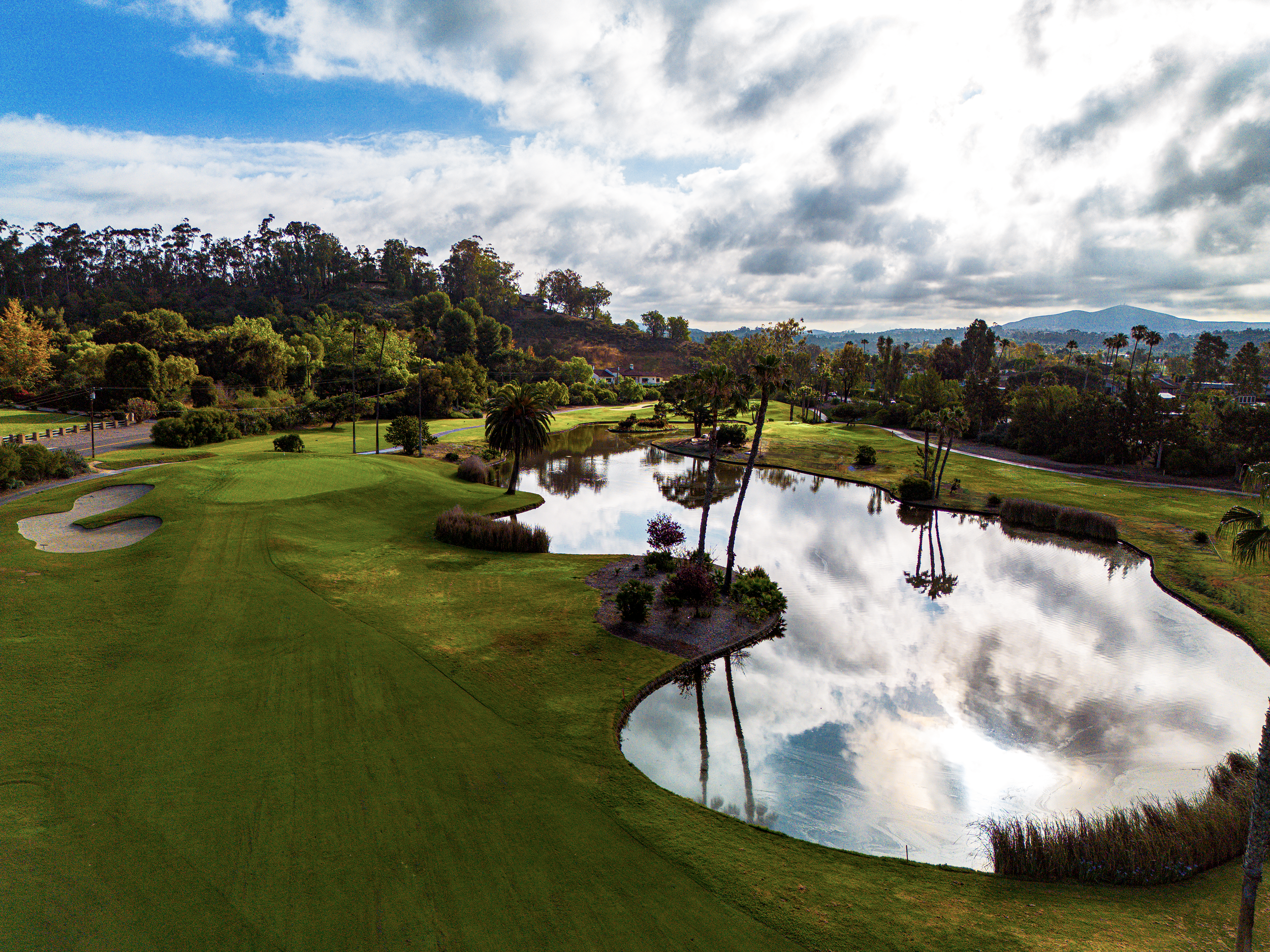A scenic landscape featuring a golf course with a small pond, surrounded by green trees and hills, under a partly cloudy sky.
