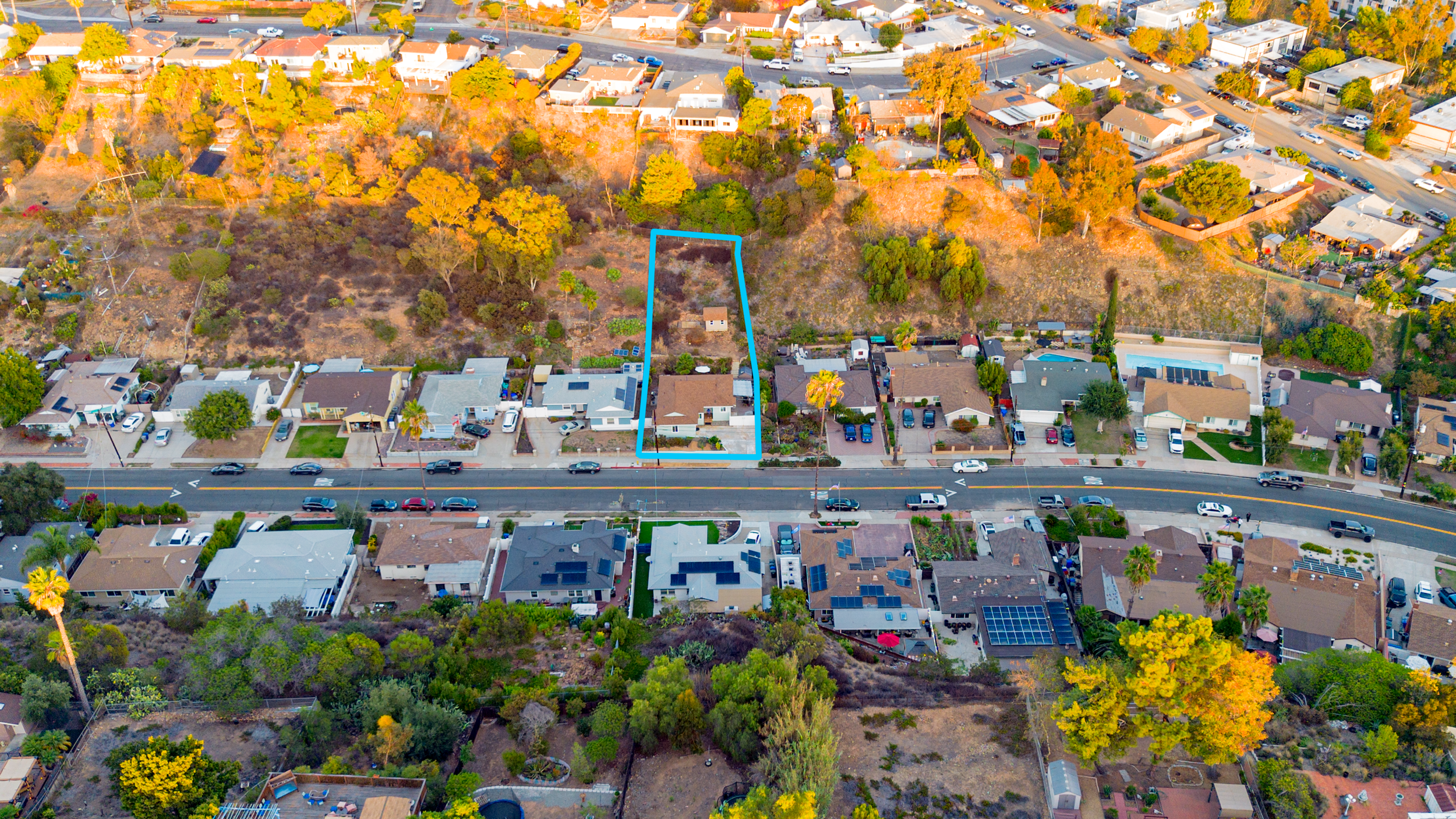Aerial view of a suburban neighborhood with a highlighted property outlined in blue, featuring a house, trees, and a backyard, with other houses and a hillside in the background.