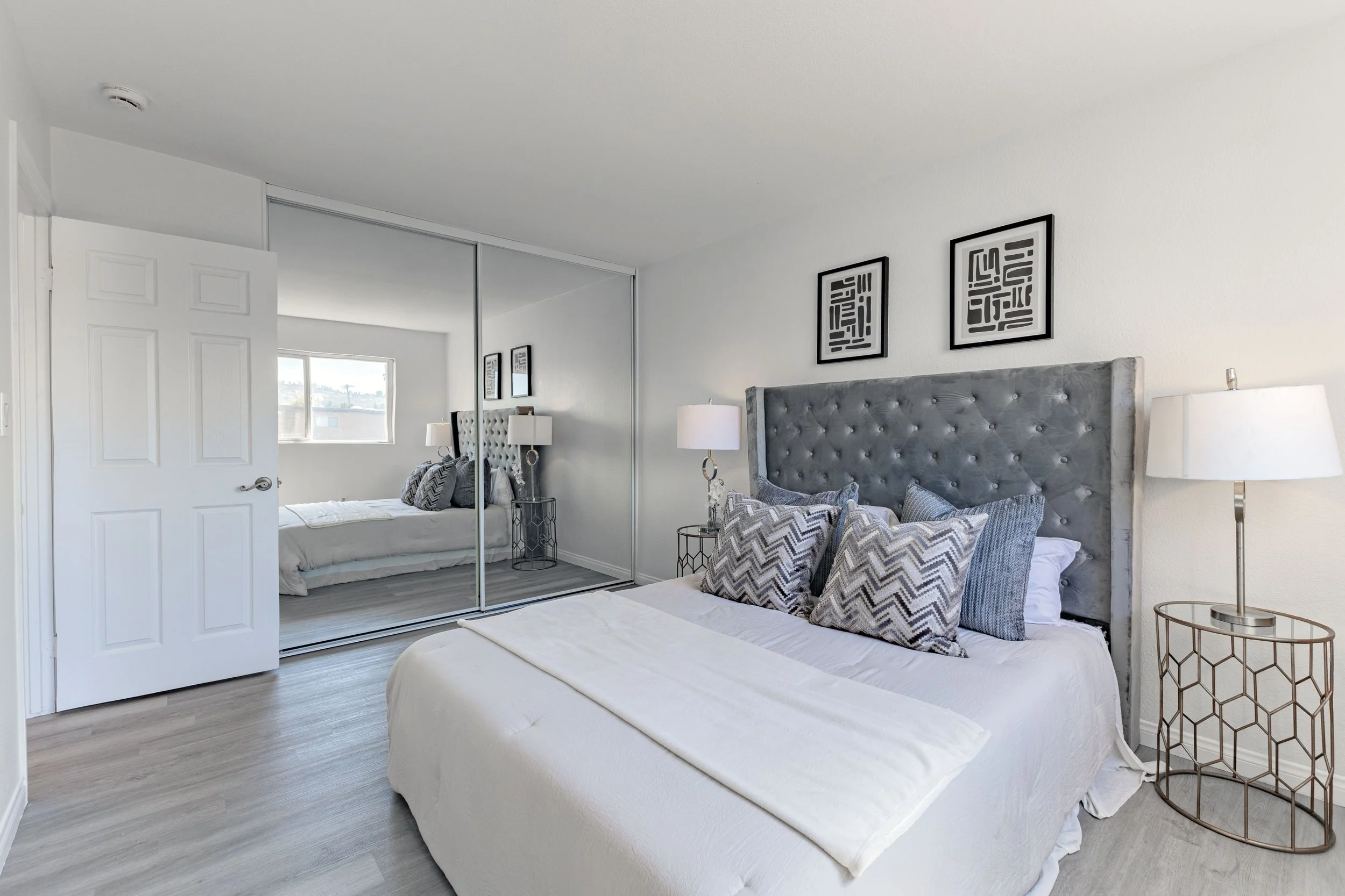 Modern bedroom with white walls, a large bed with a gray tufted headboard, multiple patterned pillows, bedside lamps, and mirrored closet doors reflecting the room.