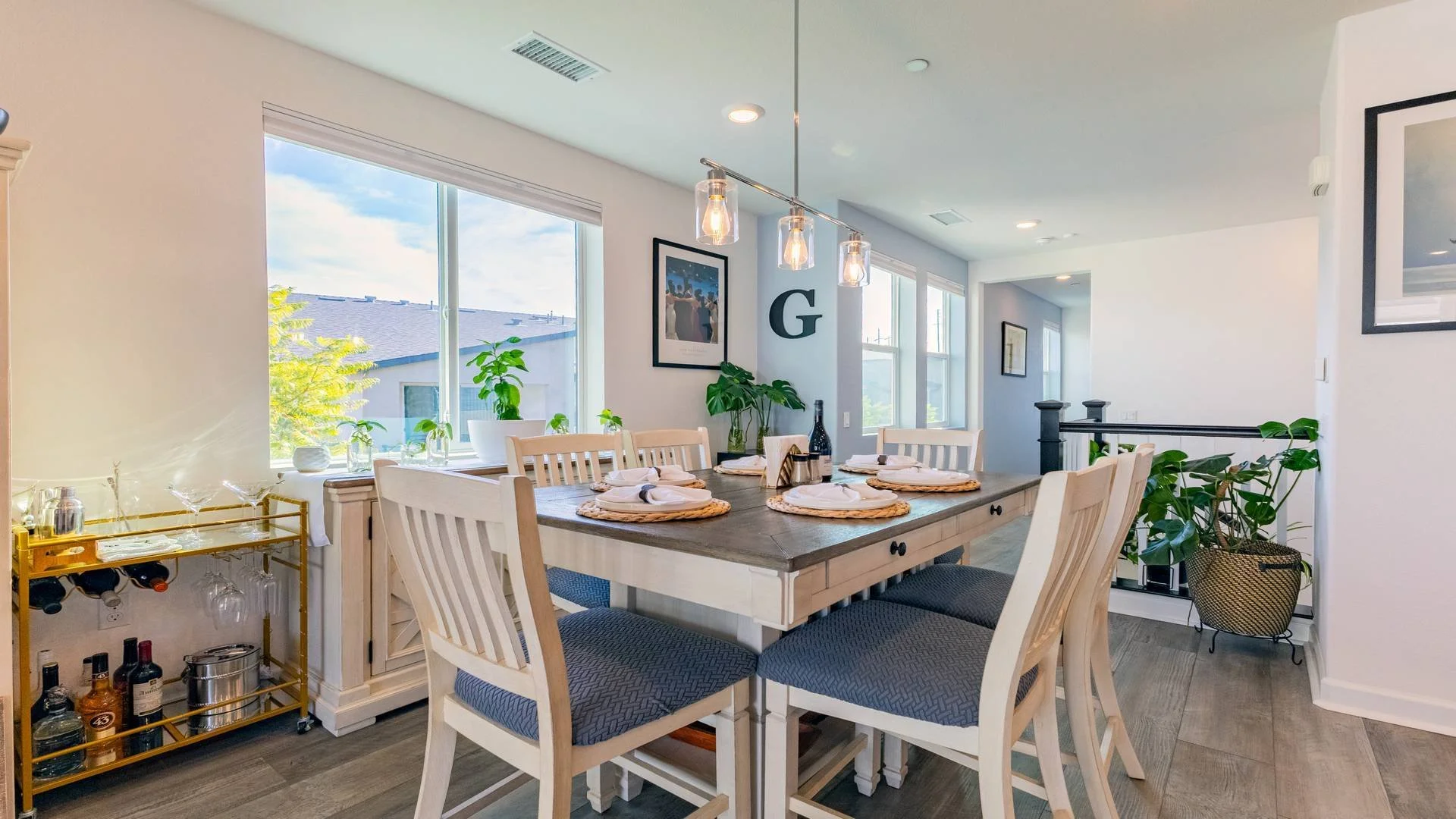Dining room with a wooden table set with cups and plates, surrounded by white chairs, large windows with a view outside, green plants, framed artwork on the wall, and a chandelier with four lights hanging above the table.