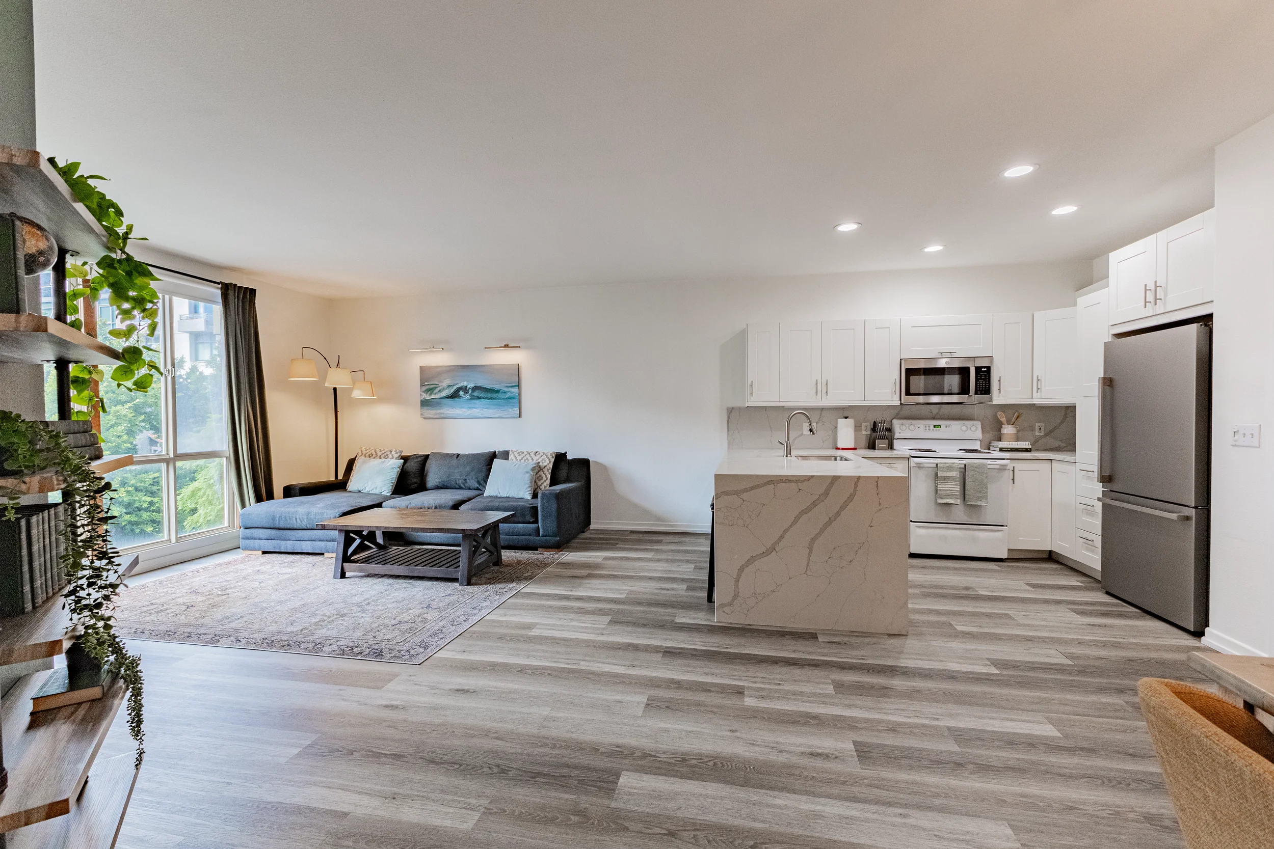 Open-concept living room and kitchen with white cabinetry, stainless steel appliances, gray flooring, a gray sofa, and large window with curtains.