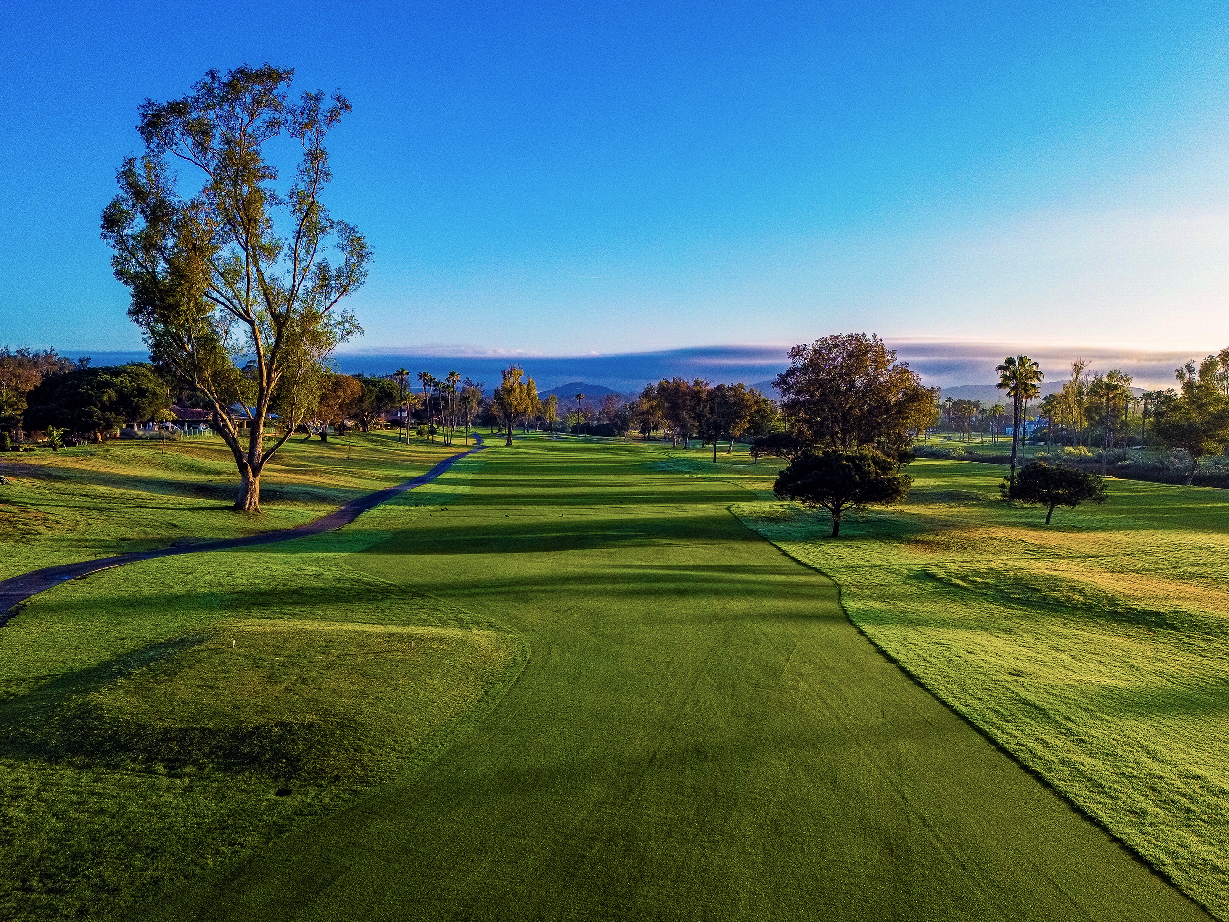 A scenic view of a golf course with green grass, trees, and a clear blue sky with some clouds.