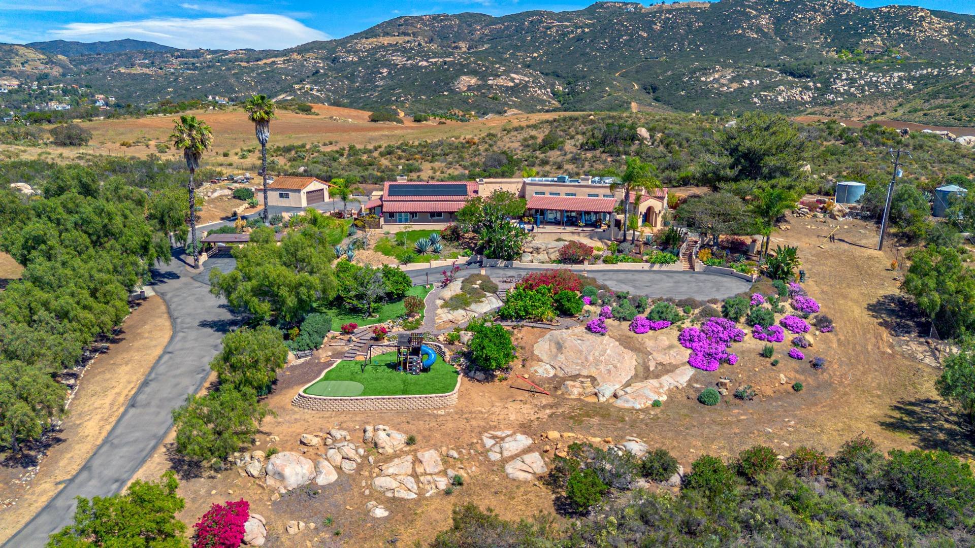 Aerial drone photography of a luxury house with a large backyard, landscaped garden with colorful flowers, trees, and a small playground. Photographed for a San Diego County real estate listing. 