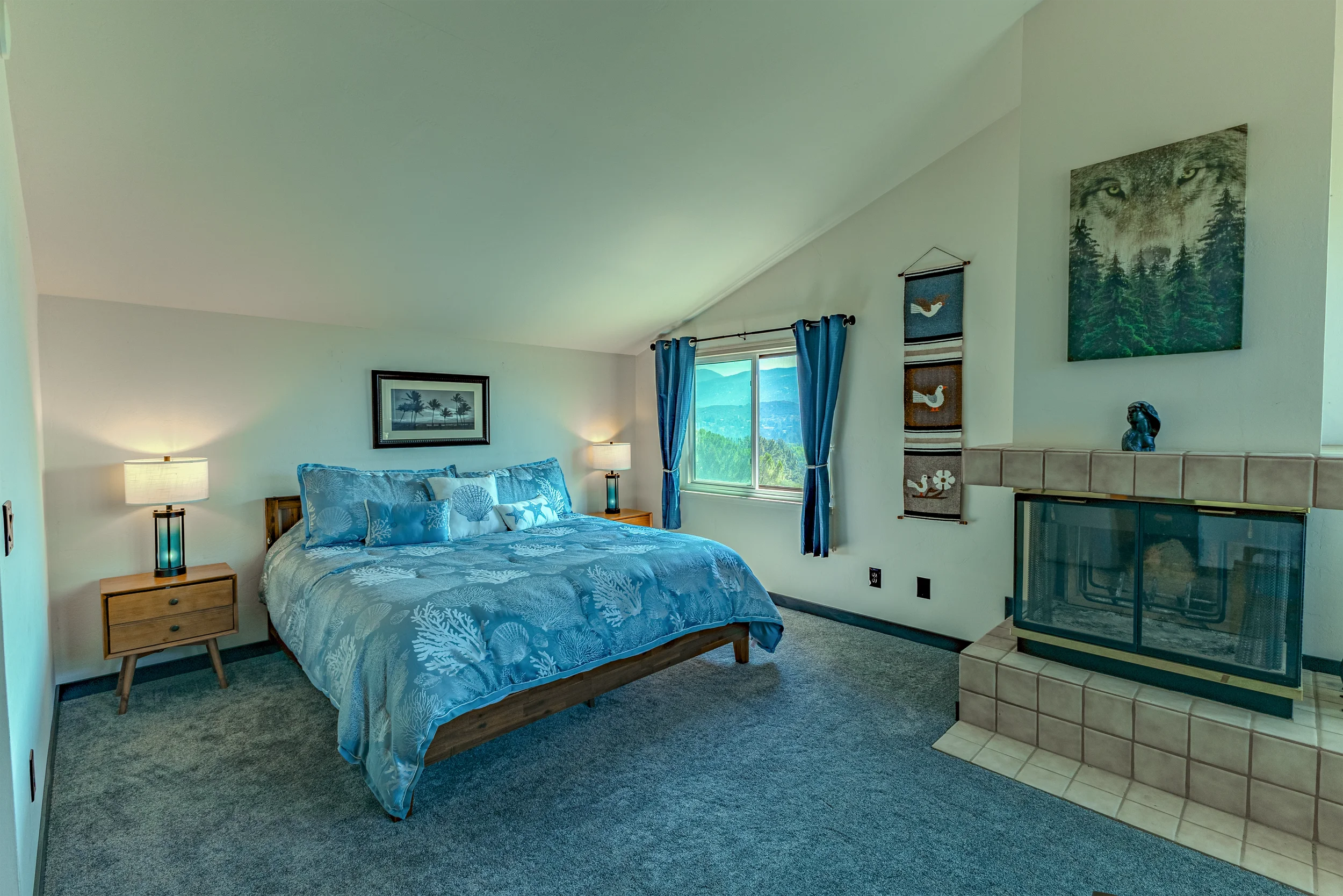 A bedroom with a bed dressed in blue sea-themed bedding, two nightstands with lamps, a window with blue curtains showing a mountain view, and artwork including a wolf portrait and bird images on the wall near a fireplace.