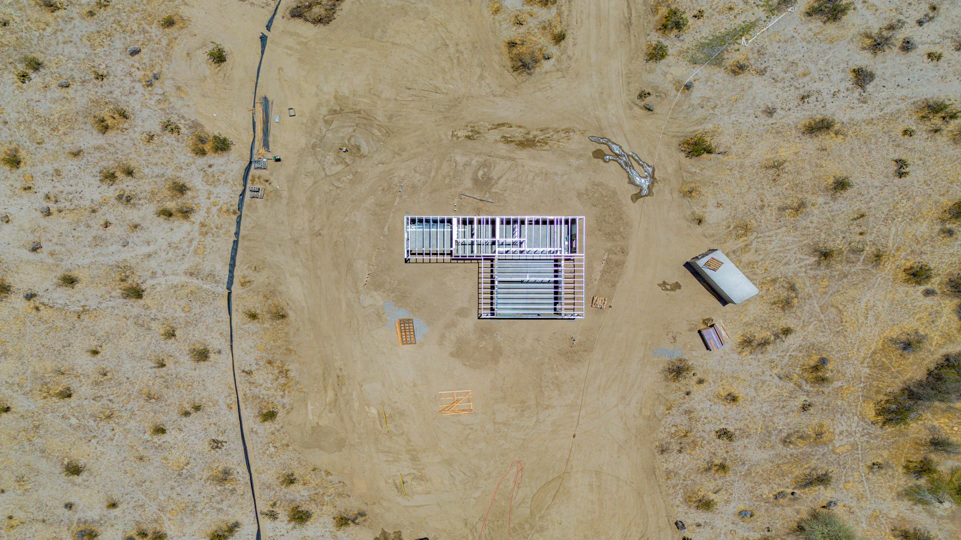 Aerial view of a house under construction in a desert area with sparse bushes, a small shed, and construction materials on the ground.