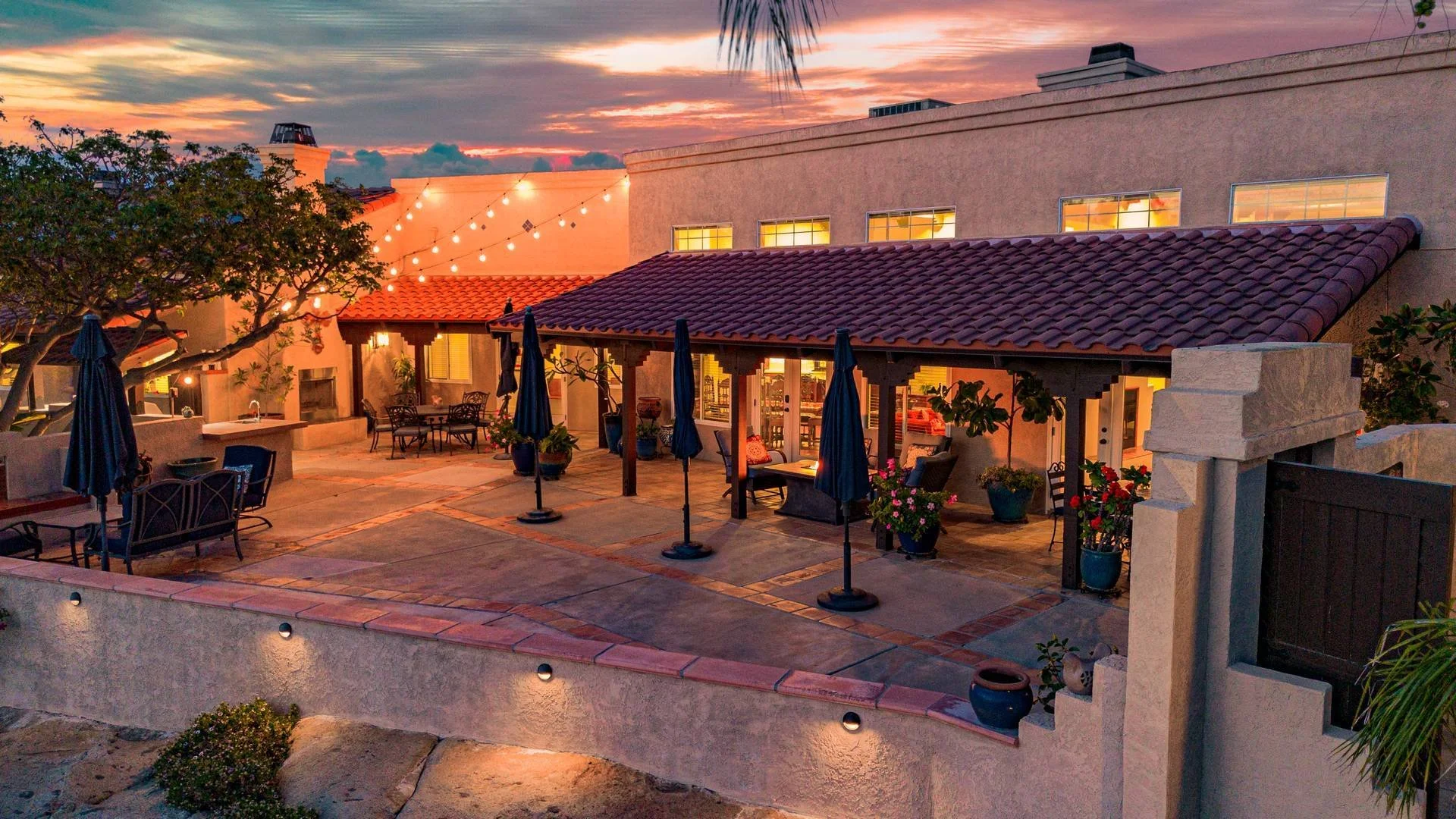 A spacious backyard patio area during sunset with a covered seating area, outdoor furniture, potted plants, string lights, and trees.