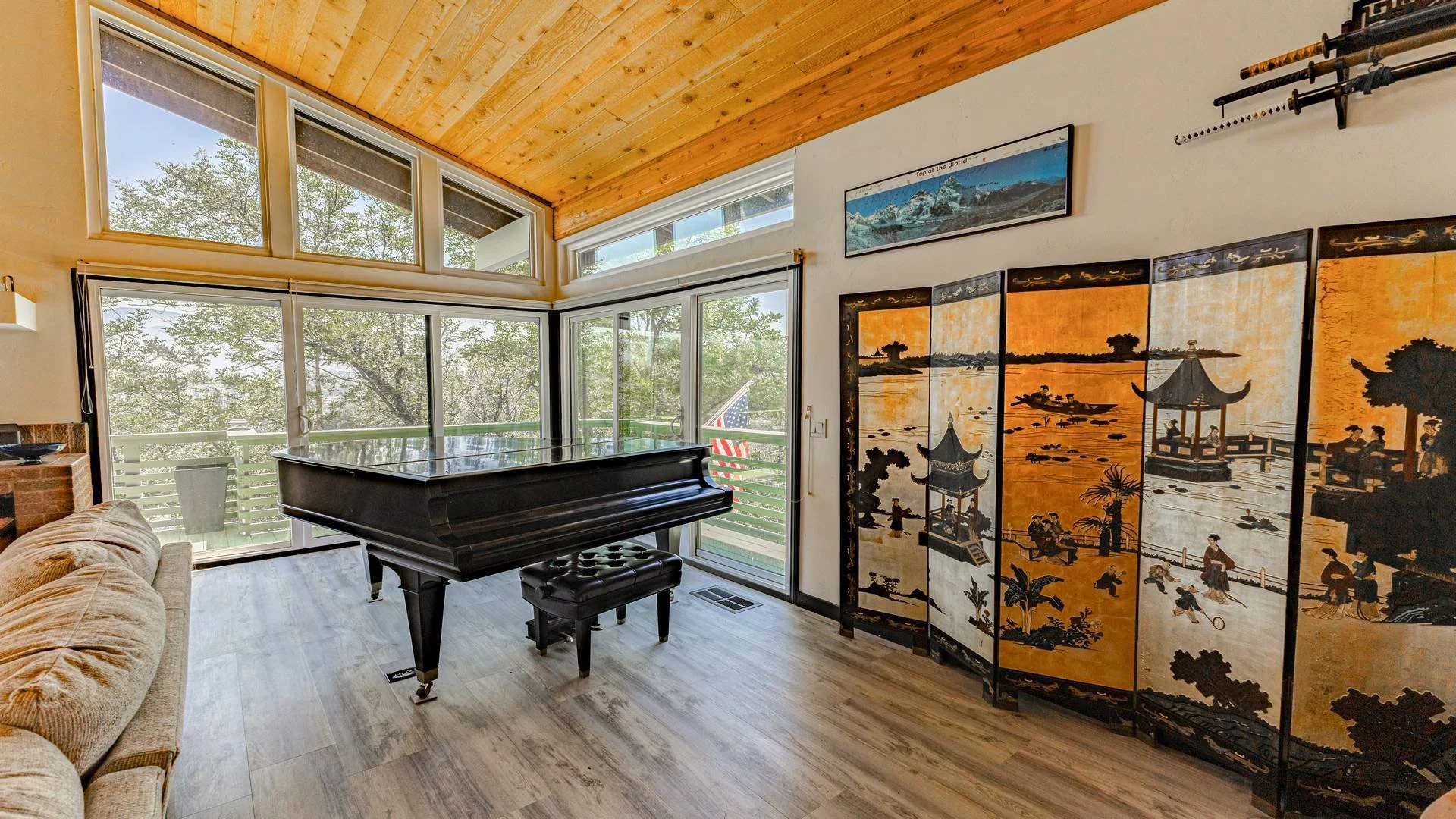 Interior of a room with large windows, a black grand piano with a matching stool, a beige sofa, and a decorative Asian folding screen with traditional artwork. The ceiling is wooden, and there is a framed panoramic photograph on the wall.