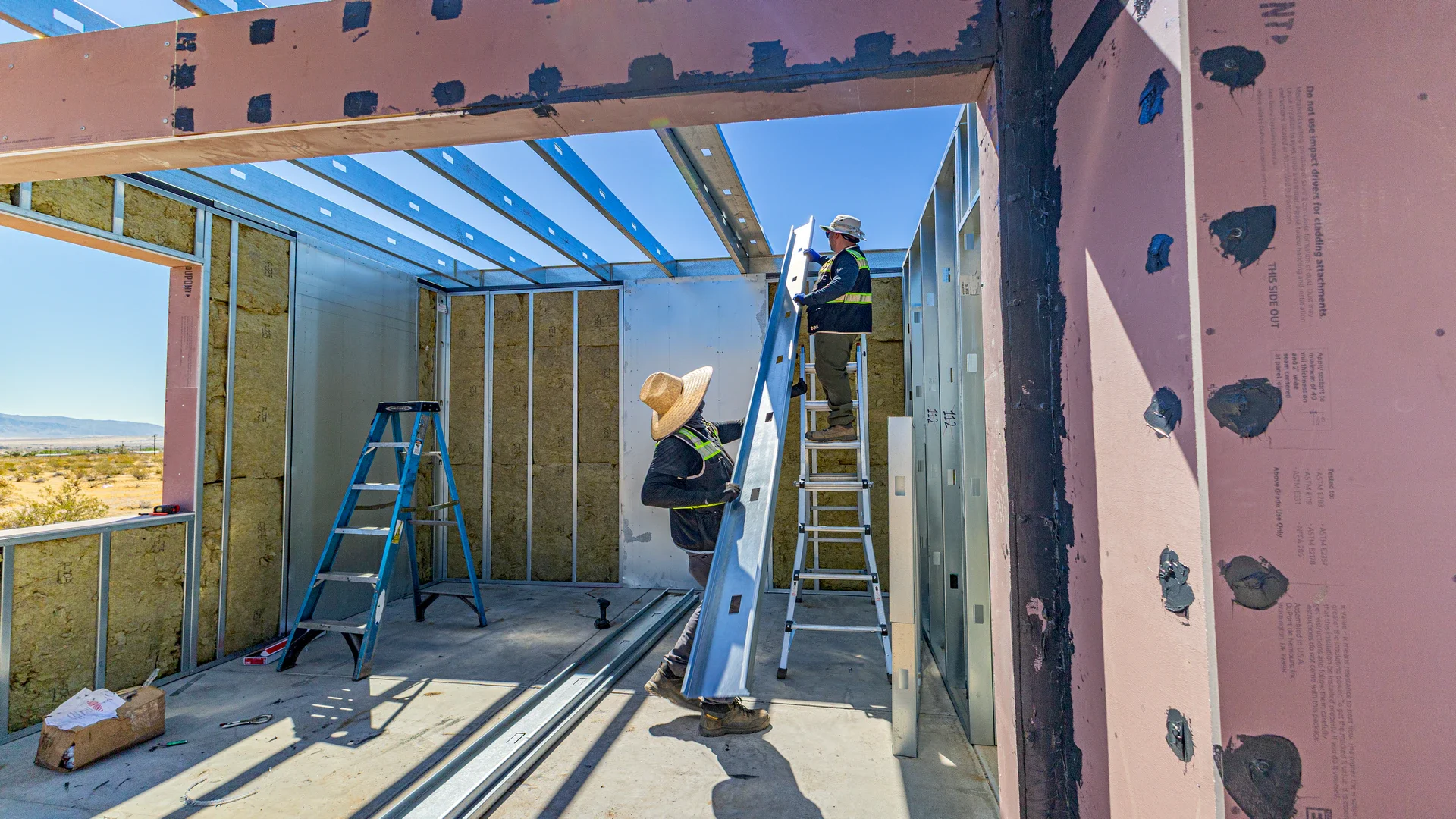 Construction workers installing metal framing inside a building under construction in a desert landscape with mountains in the distance.