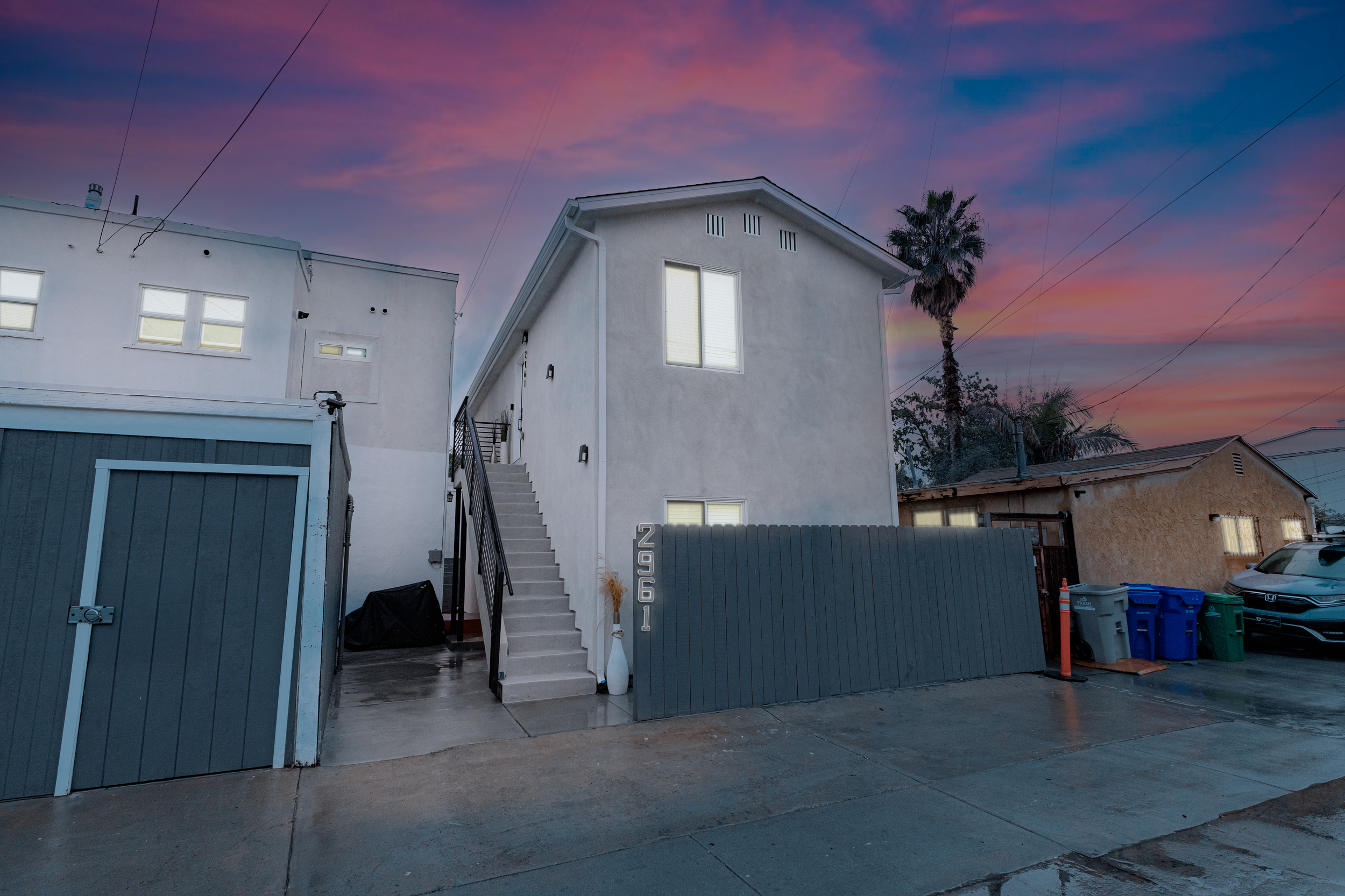 A white two-story house with exterior stairs and a dark gray fence at sunset, with palm trees and a colorful sky in the background.