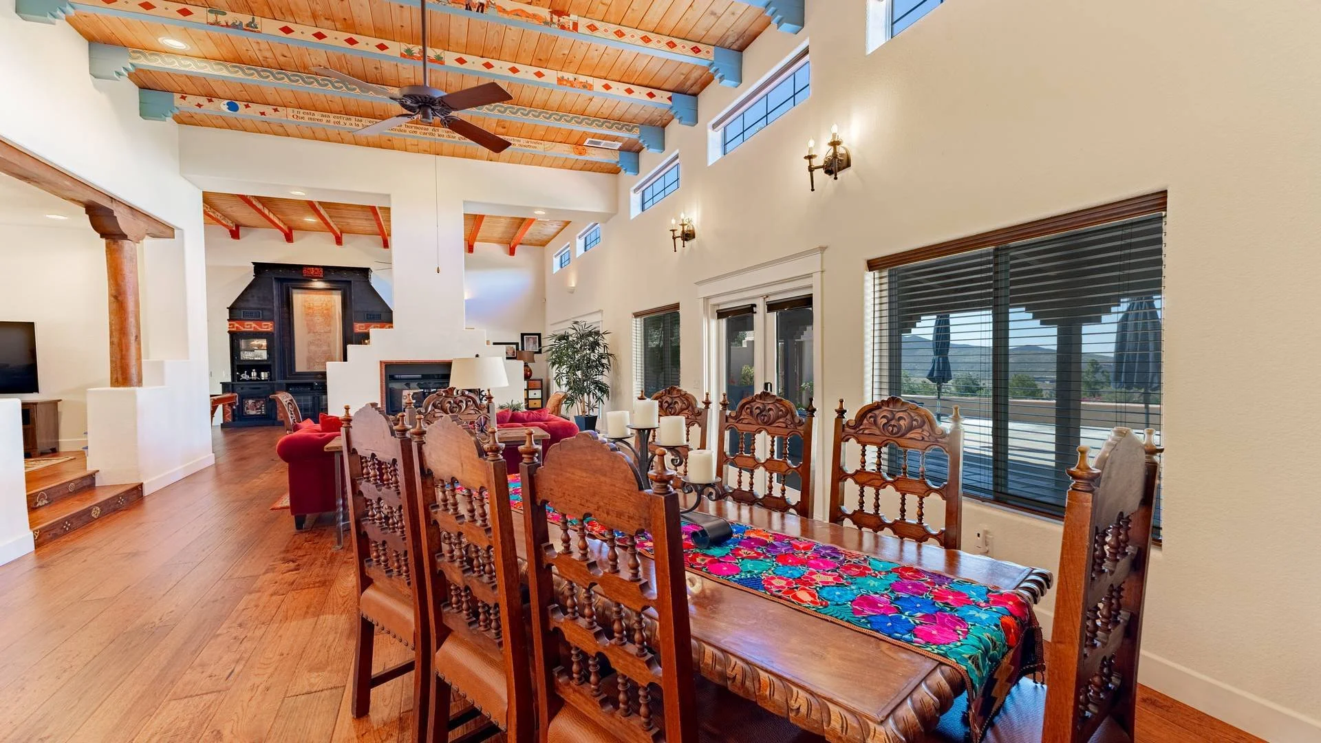 Spacious living and dining area with a high wooden ceiling, large windows, and a view of mountains. Contains a wooden dining table with a colorful tablecloth, several ornate wooden chairs, a red sofa, a fireplace, and strategic lighting fixtures.