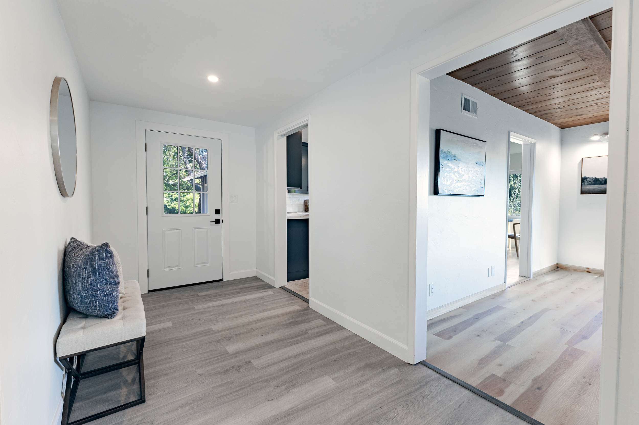 Bright entryway with a small bench, decorative pillow, and a round mirror, leading to a hallway and a room with wood ceiling.