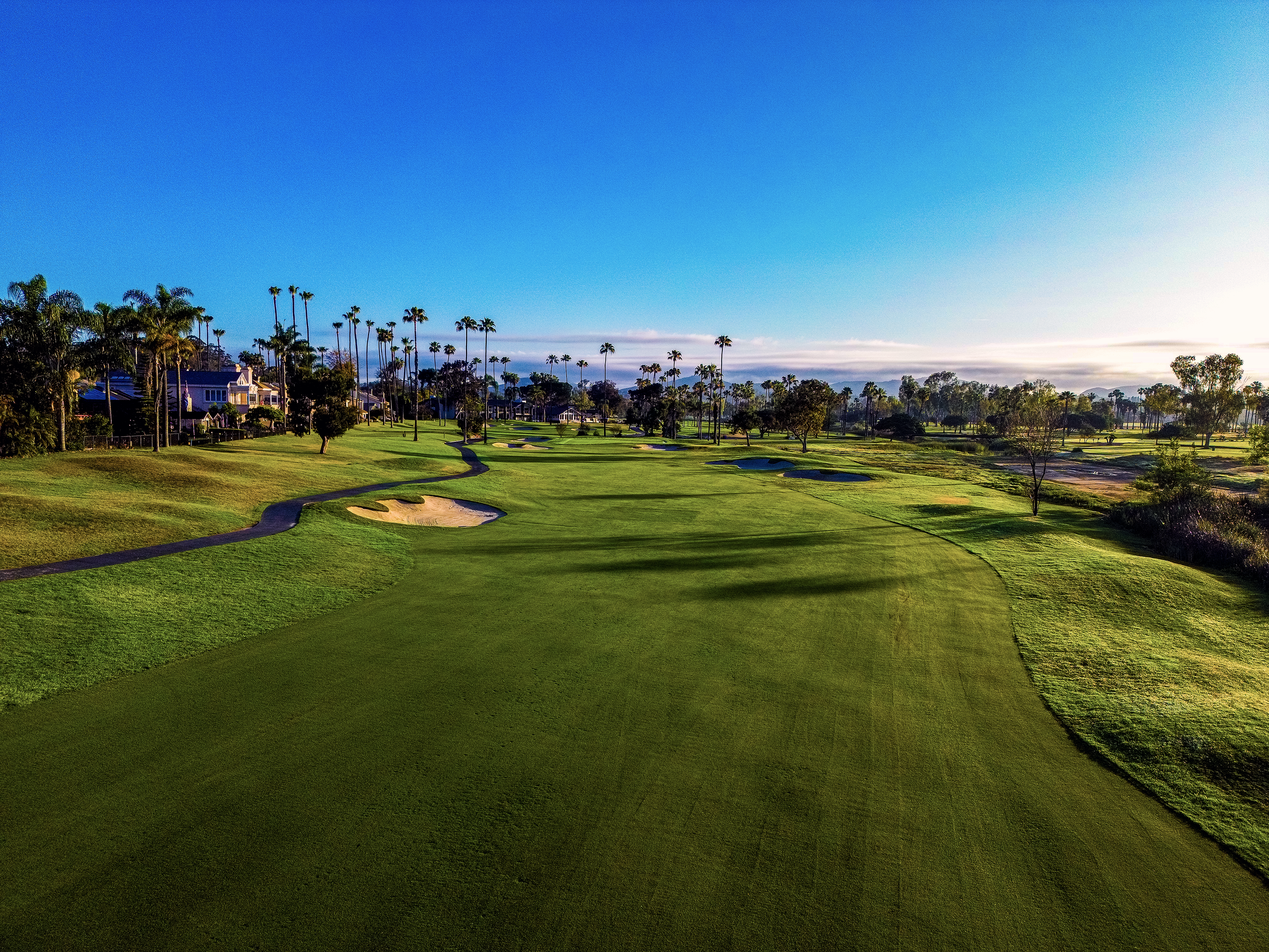 A lush green golf course with sand bunkers, surrounded by palm trees, houses, and a clear blue sky.
