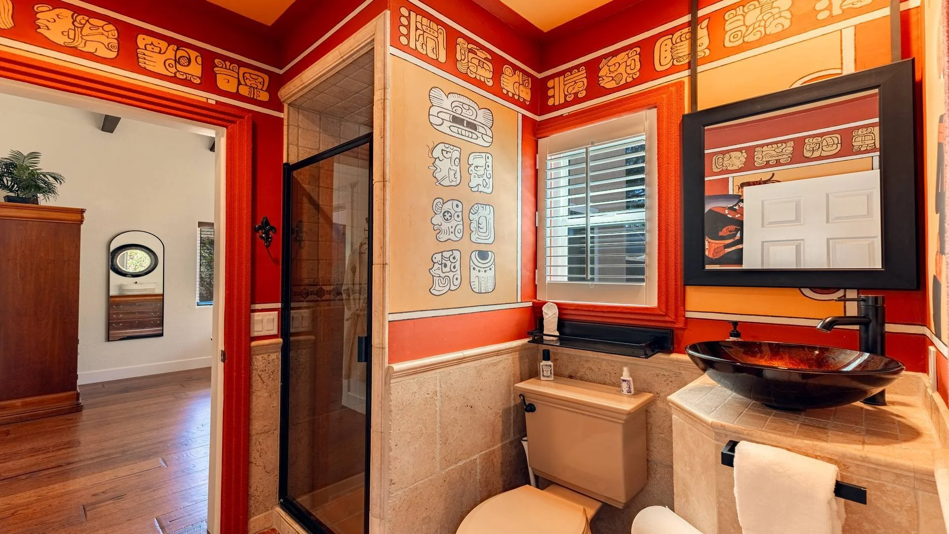 Bathroom with red and beige walls decorated with Aztec-style artwork, a black vessel sink with a modern faucet, a large mirror with a black frame, a window with white blinds, a small table with toiletries, and a shower stall with a glass door.