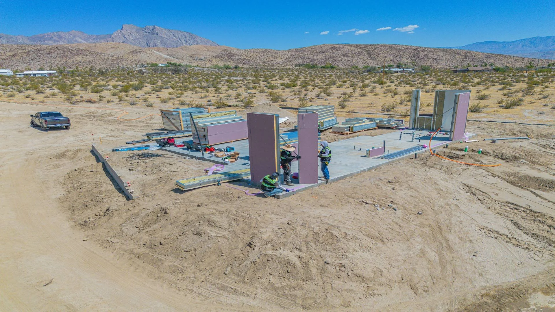 Construction site in a desert area with workers assembling building walls, with mountains in the background and a clear blue sky.