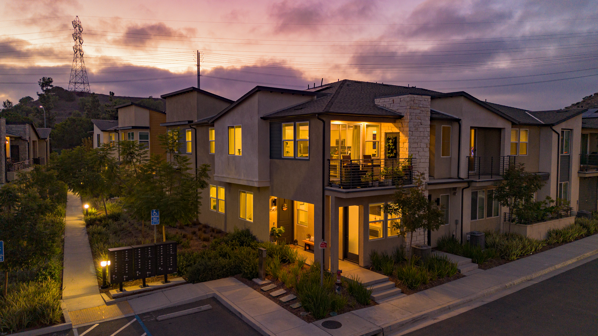 A modern apartment building at dusk with interior lights on, surrounded by landscaped greenery and a sidewalk, with hills and utility poles in the background.