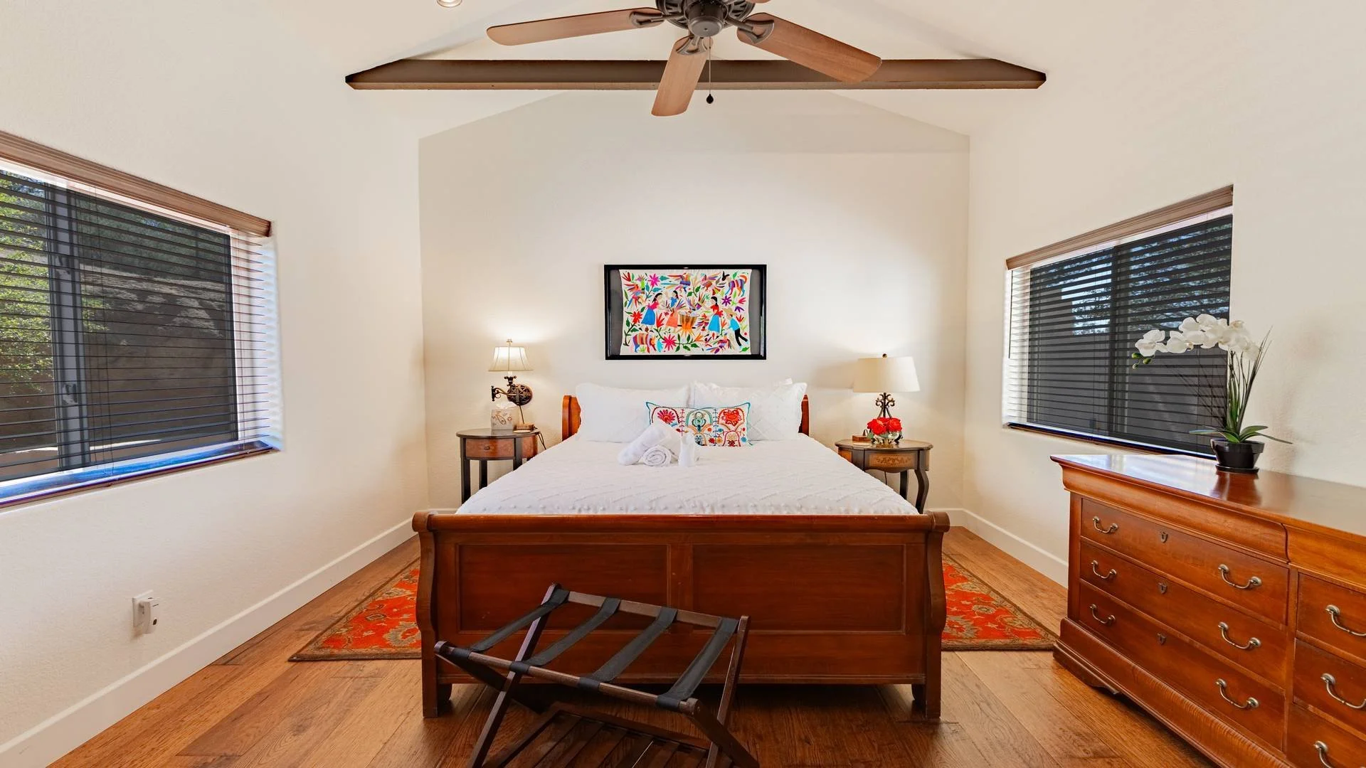 A bedroom with a wooden bed, white bedding, and colorful artwork above the headboard. Two nightstands with lamps and decorative items flank the bed. Two windows with dark blinds, a wooden dresser with a potted orchid, and a ceiling fan are also visib