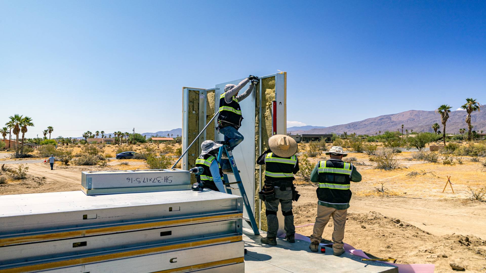 Workers in safety vests install a wall panel in a desert environment with palm trees and mountains in the background.