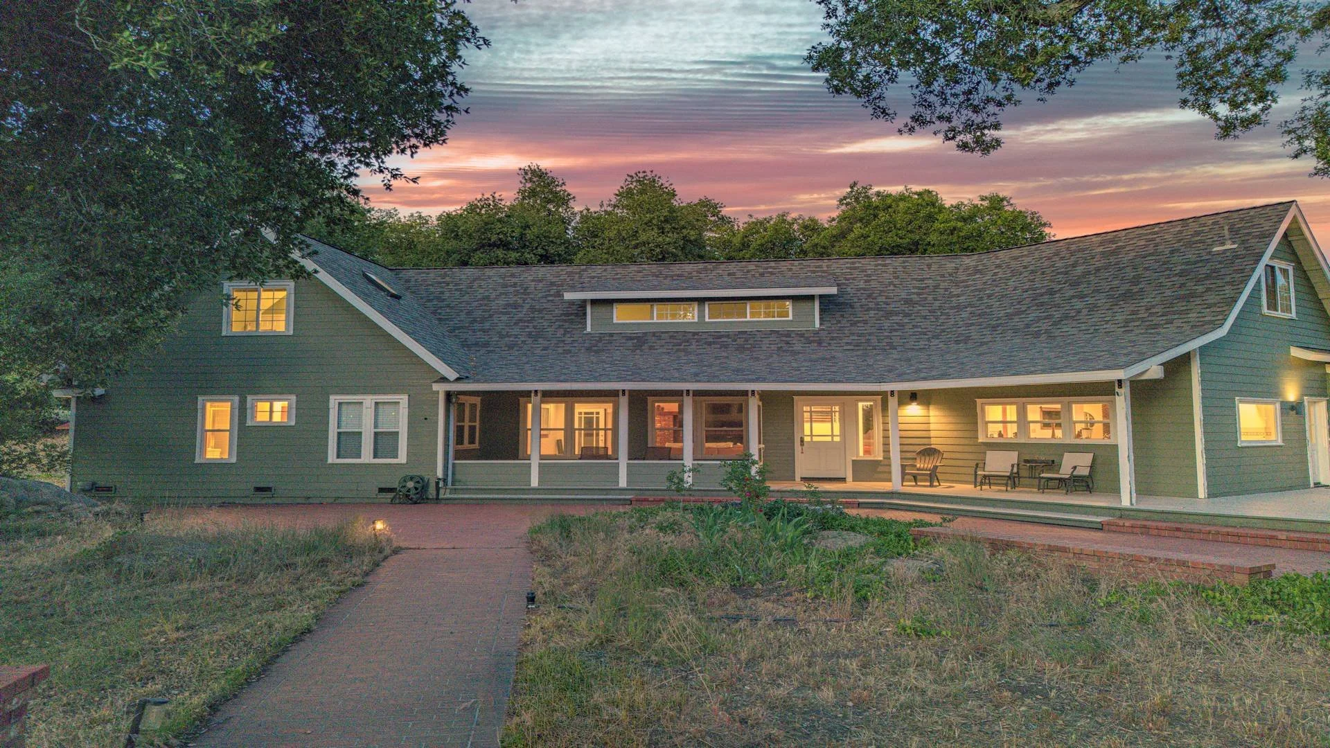 A two-story house with green siding and a dark gray roof during sunset. The house has several lit windows, a screened porch with outdoor furniture, and a brick pathway leading to it. Trees surround the house, and the sky features colorful clouds.