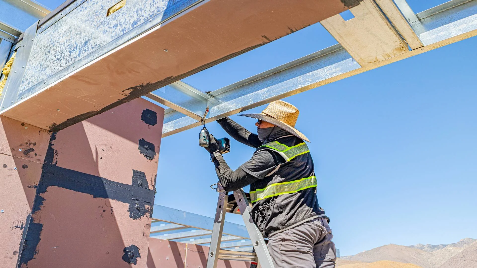 A construction worker wearing a wide-brimmed straw hat, sunglasses, a face mask, and a reflective safety vest, standing on a ladder, installing or repairing metal framing on the exterior of a building against a clear blue sky.