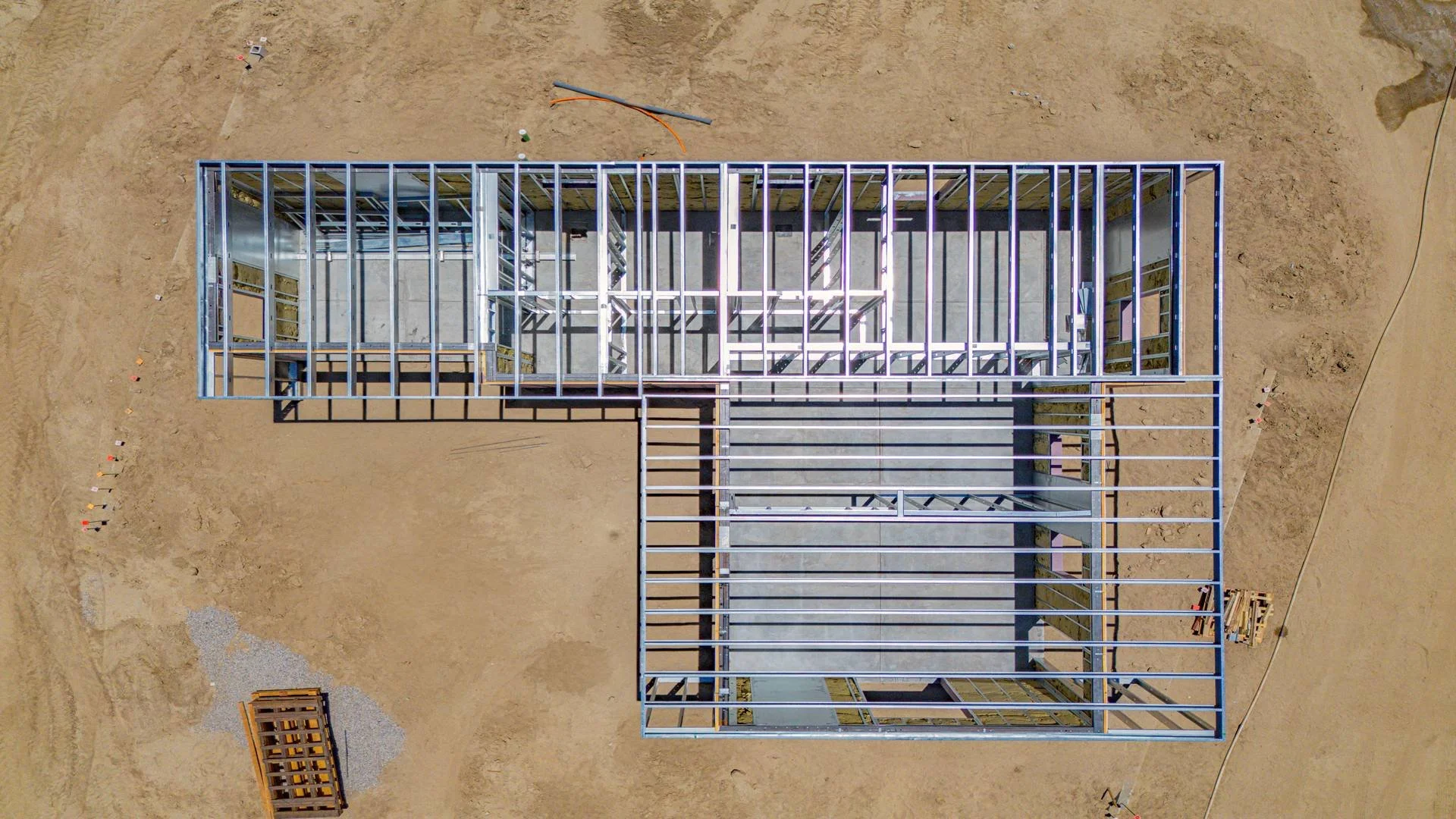 An aerial view of a partially constructed building frame on a dirt lot, with metal beams and concrete flooring. Some building materials are visible around the site.