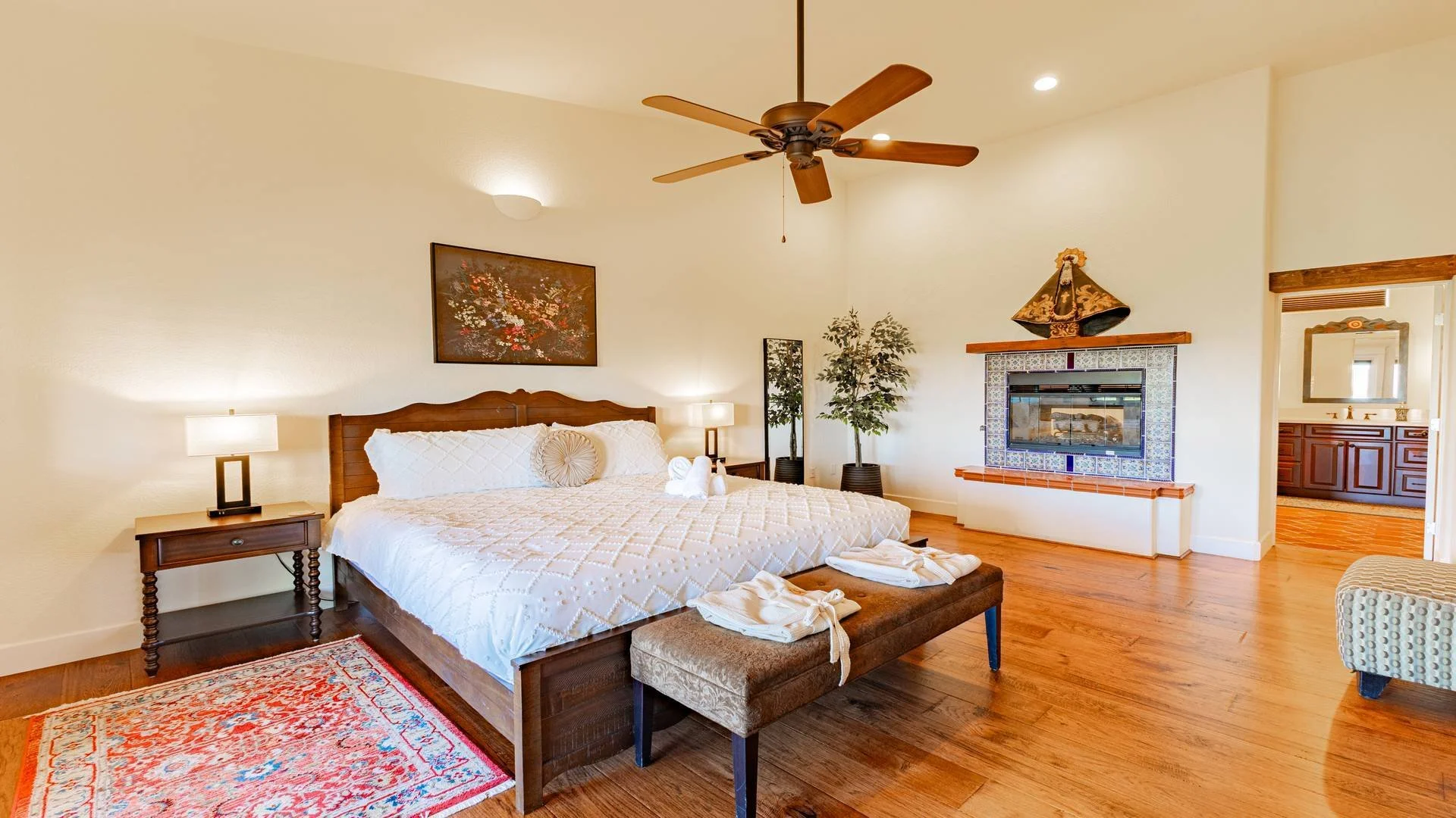 A bedroom with a wooden bed, white bedding, and a decorative round pillow. There are two bedside tables with lamps, a painting above the bed, a ceiling fan, a fireplace with decorative tiles, a mirror in the bathroom, and a potted plant. The room has