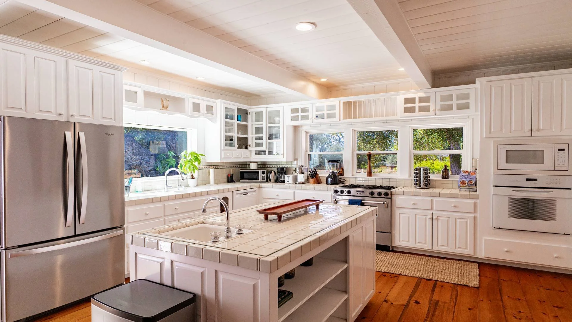 Luxury photography of a  bright white kitchen with large windows, stainless steel refrigerator, white cabinets, tiled countertops, and wooden flooring. Taken for an Airbnb short-term rental listing in San Diego County. 