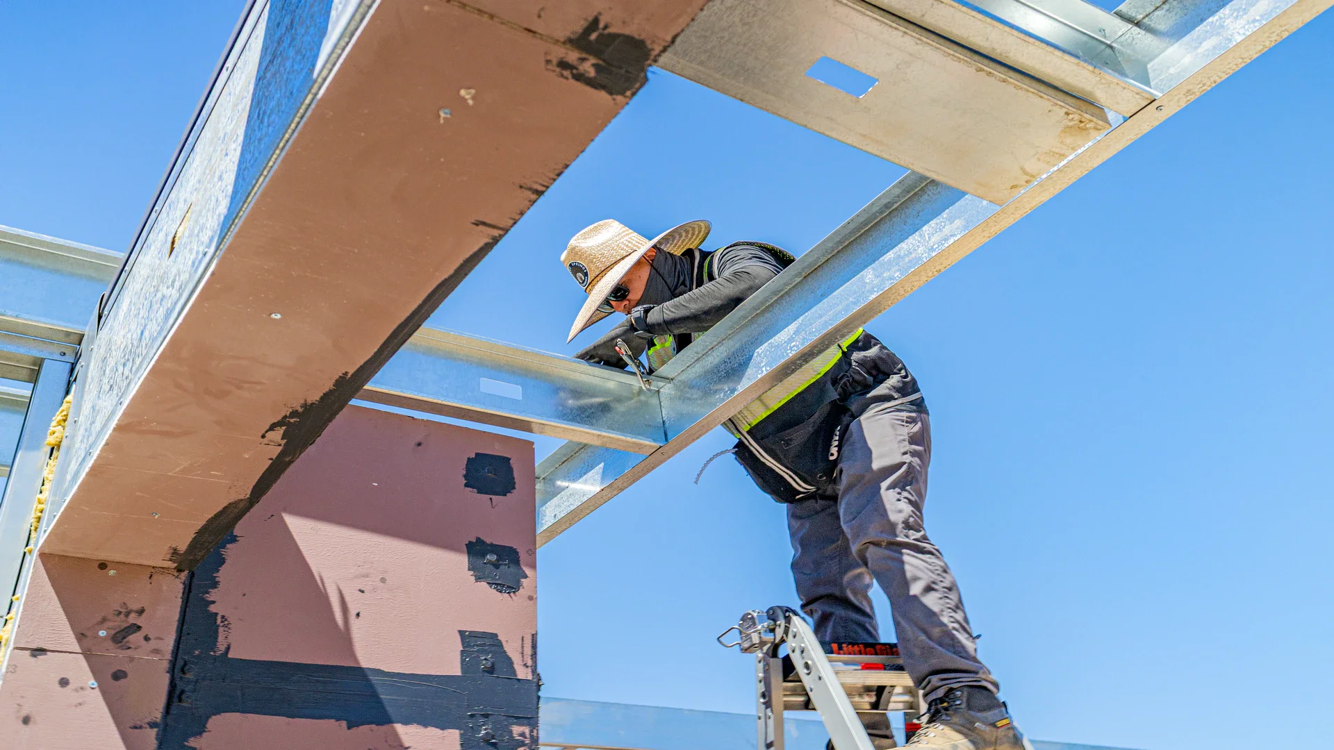 A construction worker wearing a wide-brimmed hat, sunglasses, a face mask, and work gloves, working on a metal structure at a building site under a clear blue sky. The worker is standing on a ladder and appears to be fastening or adjusting parts of the framework.