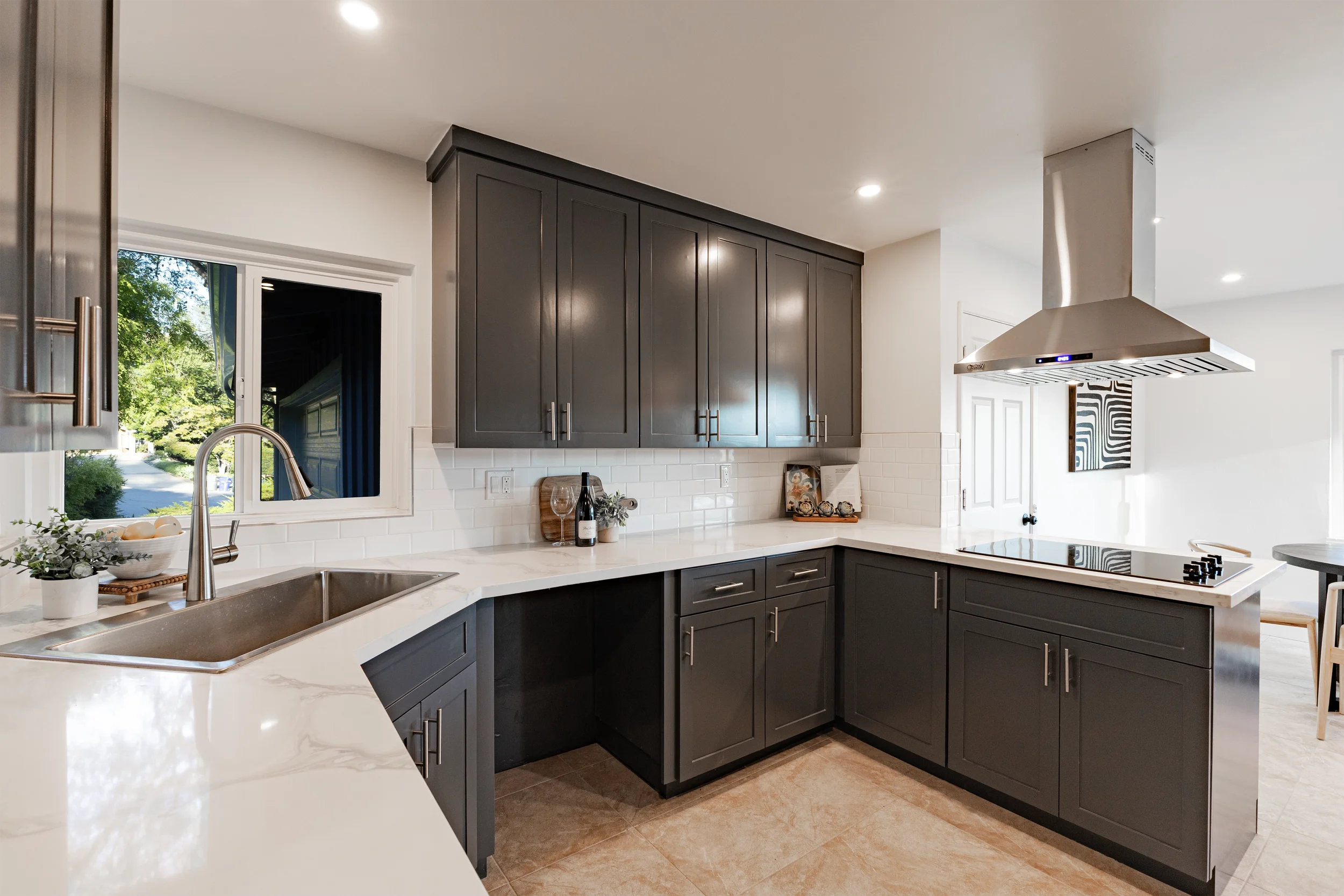 Modern kitchen with dark gray cabinets, white countertops, a stainless steel range hood, and a window overlooking a green outdoor area.