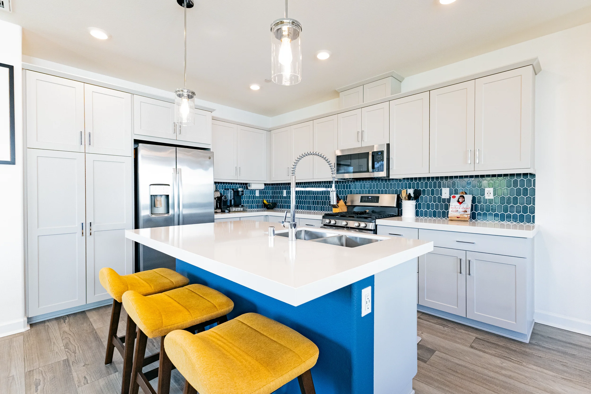 Modern kitchen with white cabinets, blue hexagonal tile backsplash, stainless steel refrigerator and oven, kitchen island with a white countertop, yellow stools, and pendant lighting.