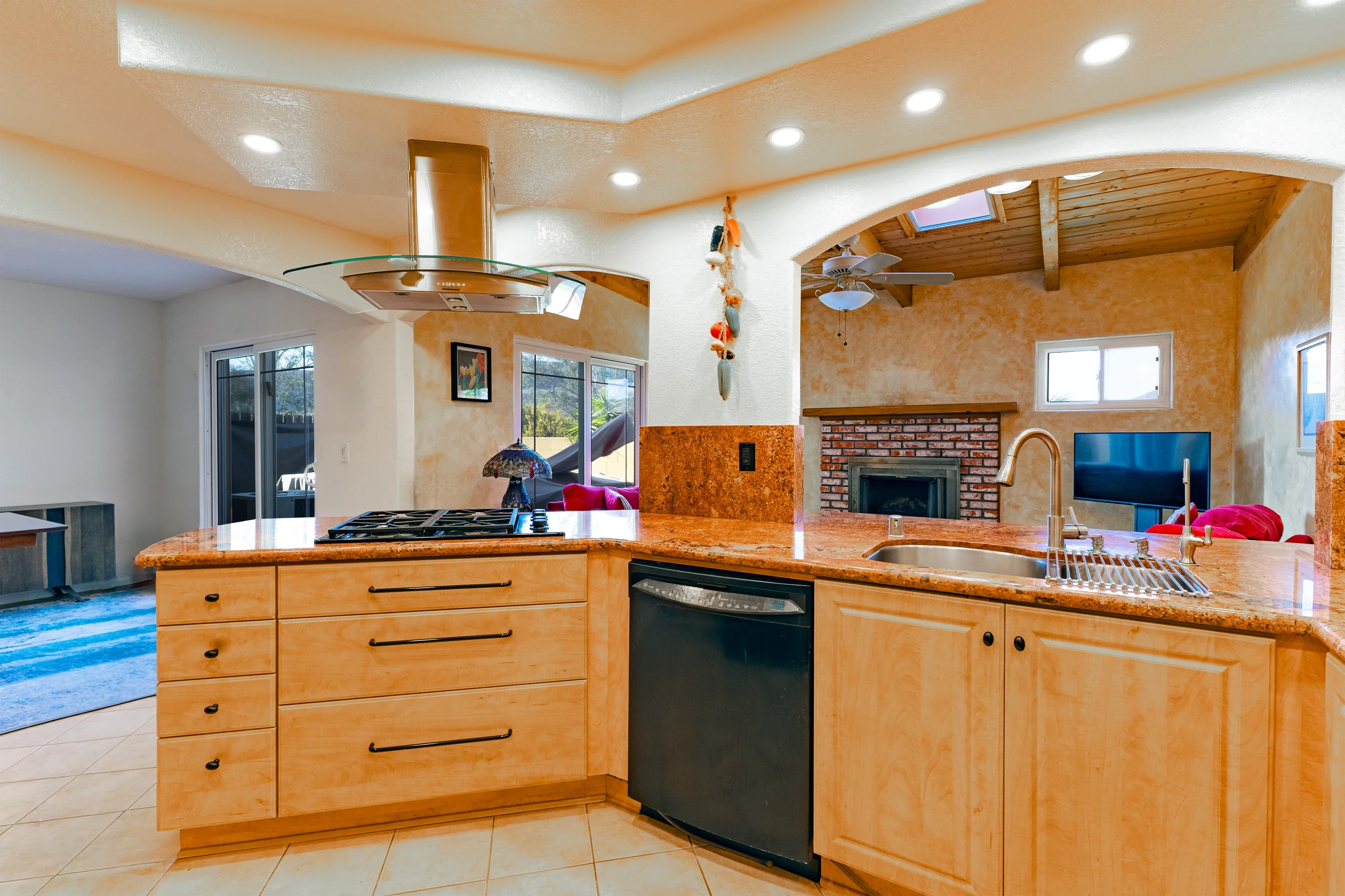 Open-plan kitchen with wooden cabinets and granite countertops, gas stove with a glass range hood, and a sink with a drying rack. In the background, a living room features a brick fireplace, a TV, and a ceiling fan, with natural light coming through 
