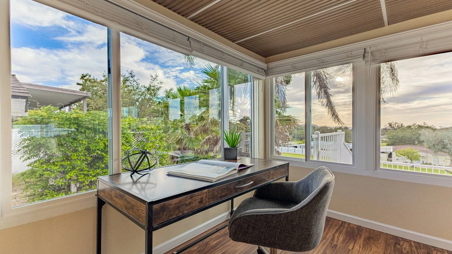 View of a sunroom with large windows showing greenery and trees outside, a wooden desk with a book, a potted plant, and a decorative sphere, and a gray office chair.