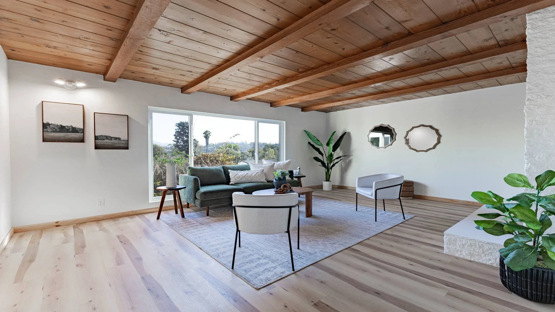 Bright living room with wooden ceiling, white walls, large window, green couch, white armchair, coffee table, plants, and framed mirrors. Real estate photographer in San Diego County. 