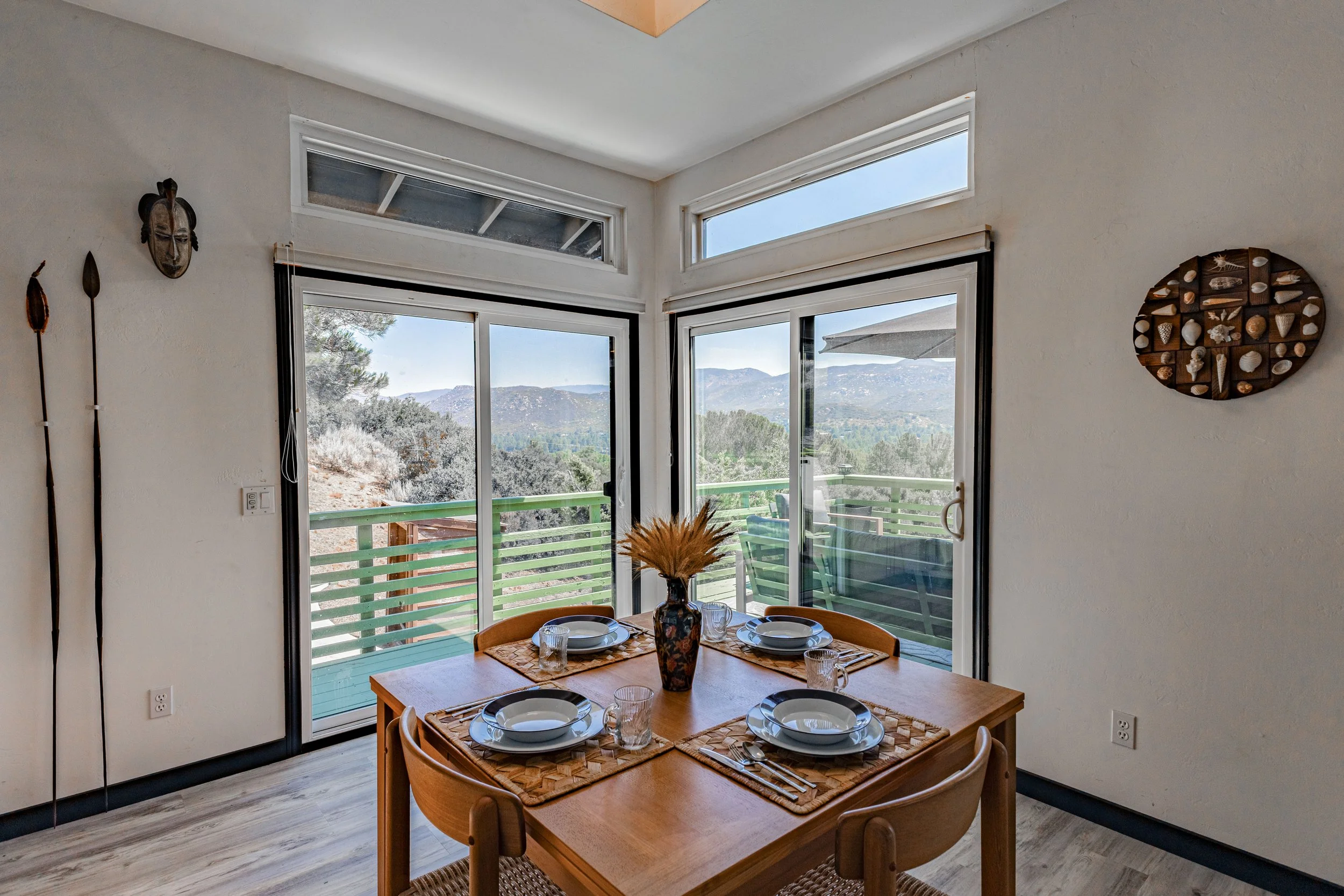 Dining area with a wooden table set for four, featuring plates, glasses, and cutlery, with a vase of dried wheat in the center, overlooking a mountain view through large glass sliding doors leading to a balcony.