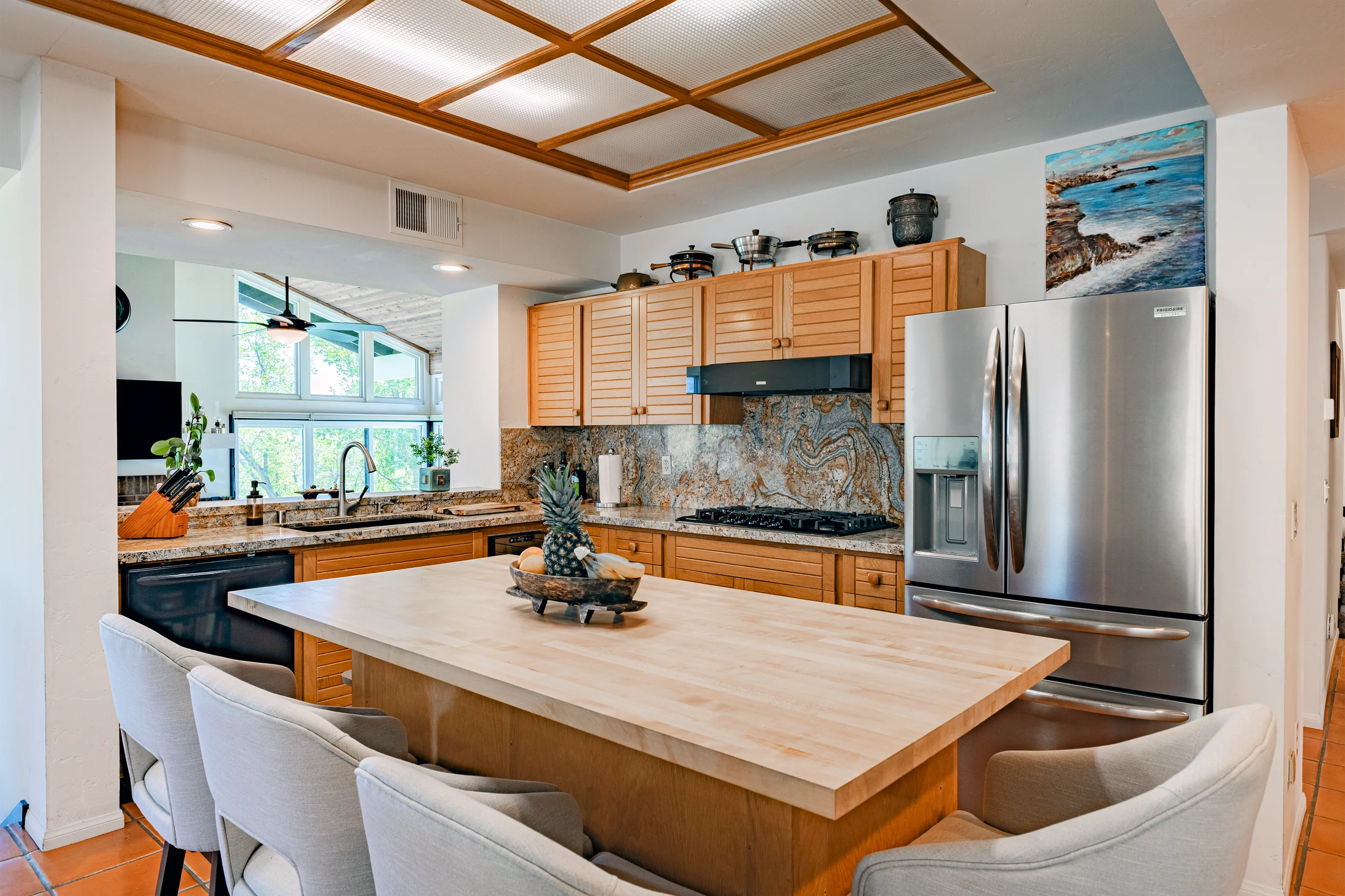Bright, modern kitchen and dining area with white cabinets, stainless steel appliances, a round marble dining table, and four white cushioned chairs. Large windows with blinds let in natural light. A chandelier hangs above the dining table, and a potted plant is in the corner.